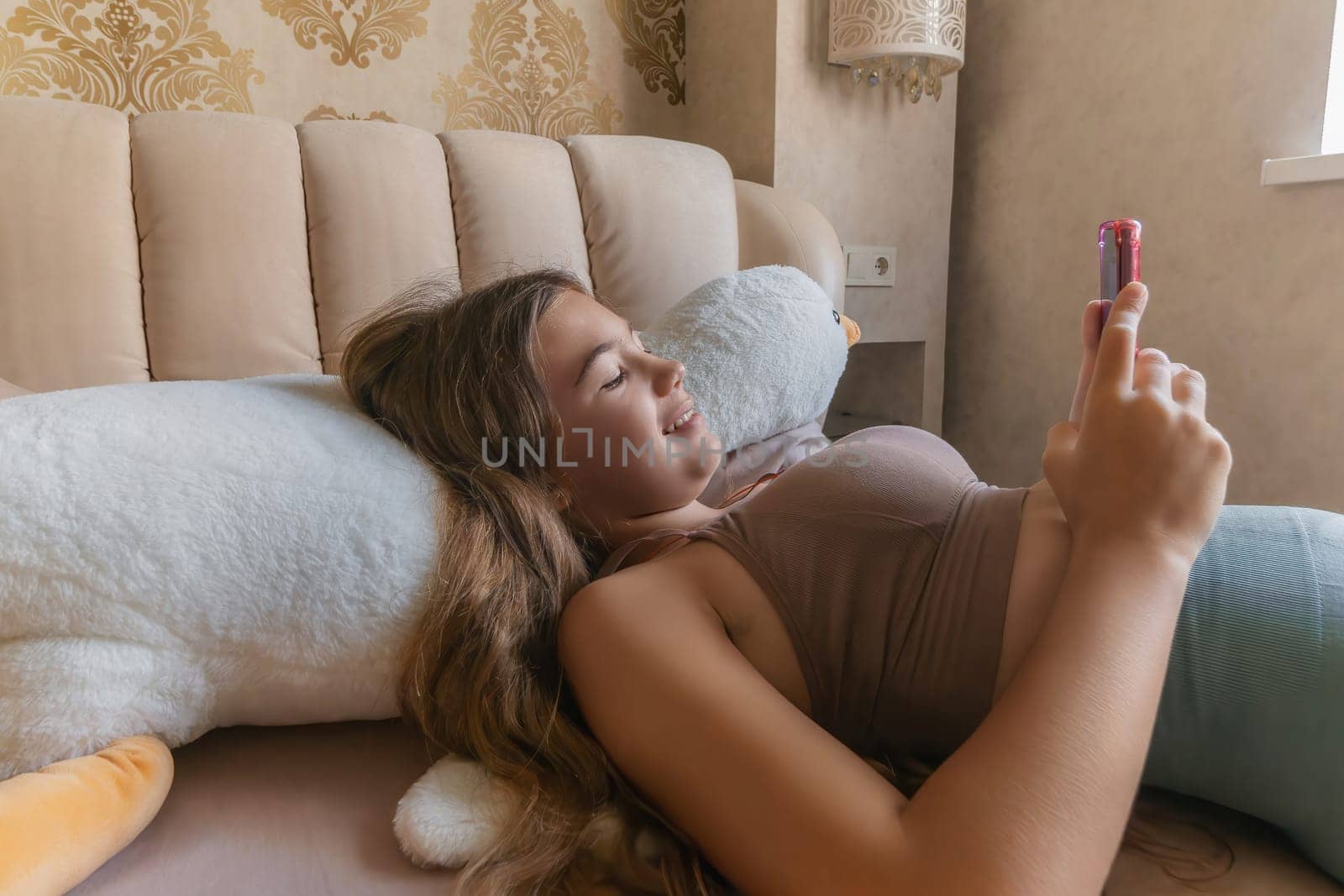 A girl is laying on a bed with a pink phone in her hand. She is looking at the phone and she is focused on something on the screen. by Matiunina