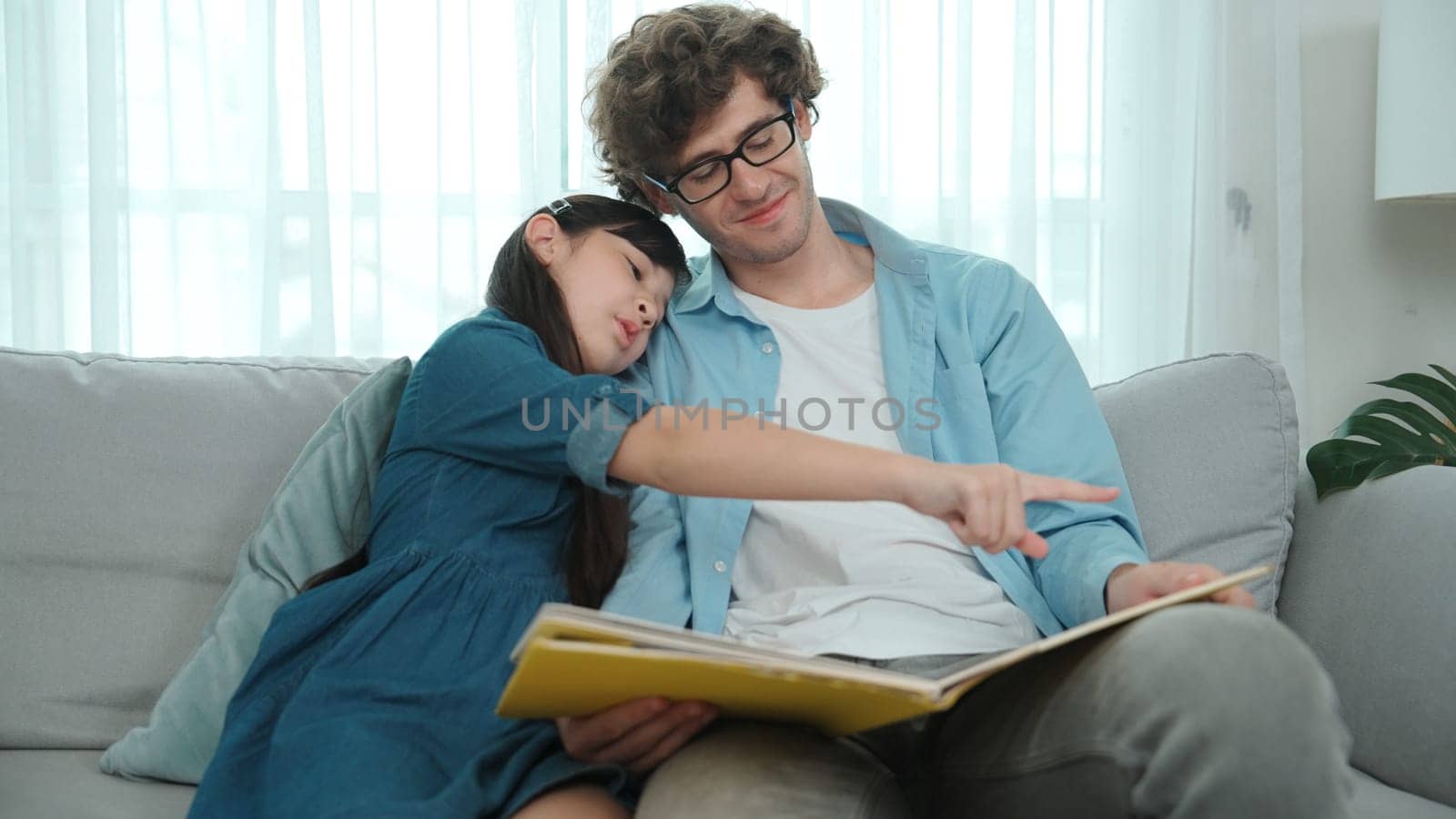 Happy daughter lean on father shoulder while listening story telling. Young asian schoolgirl looking ar dad while reading story book together and sitting at sofa. Family recreation concept. Pedagogy.