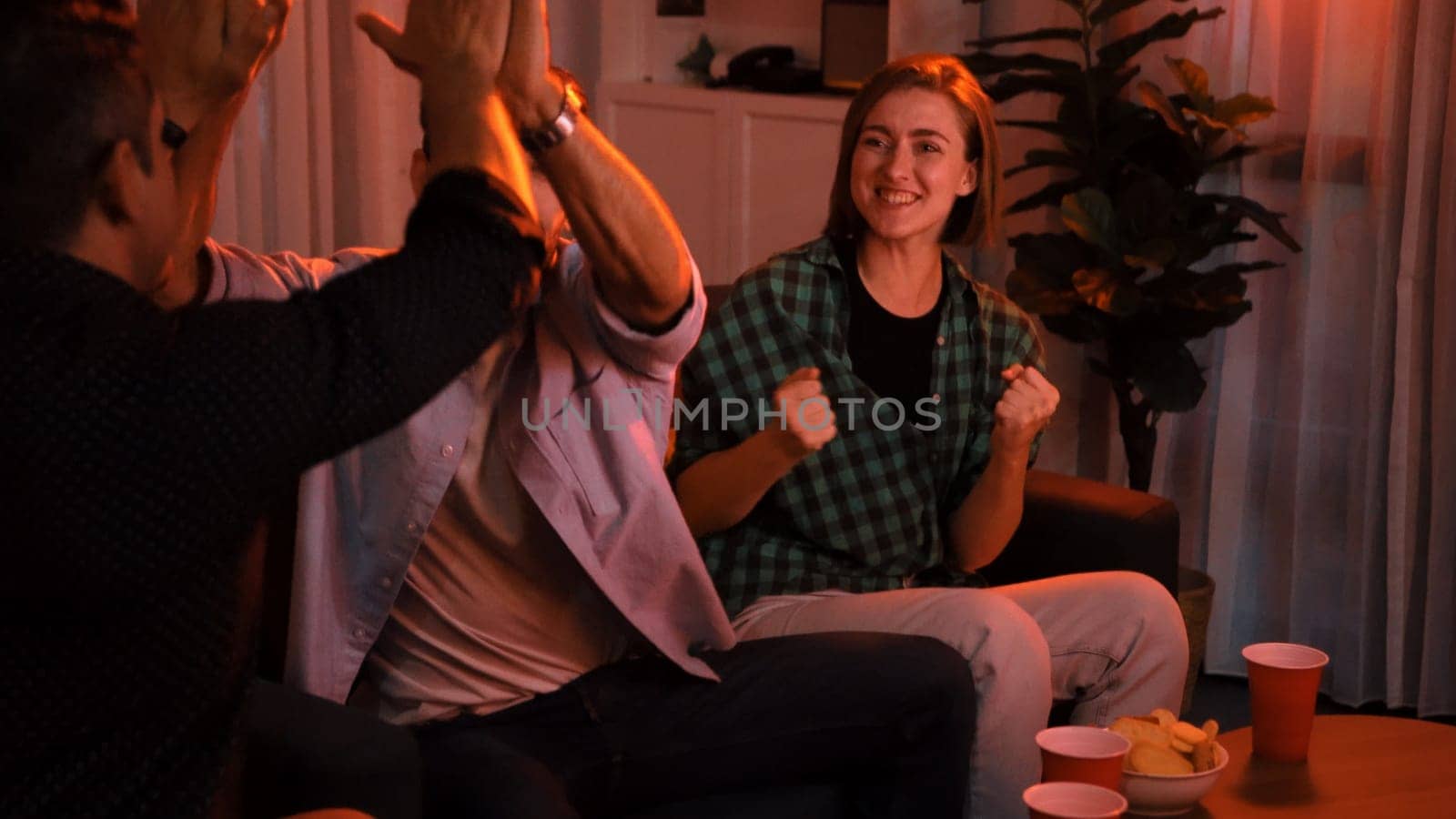 Family and colleague sitting at dark living room with red filter while cheering football team together. Caucasian people enjoy watching tv and giving high five to celebrate winning team. Convocation.