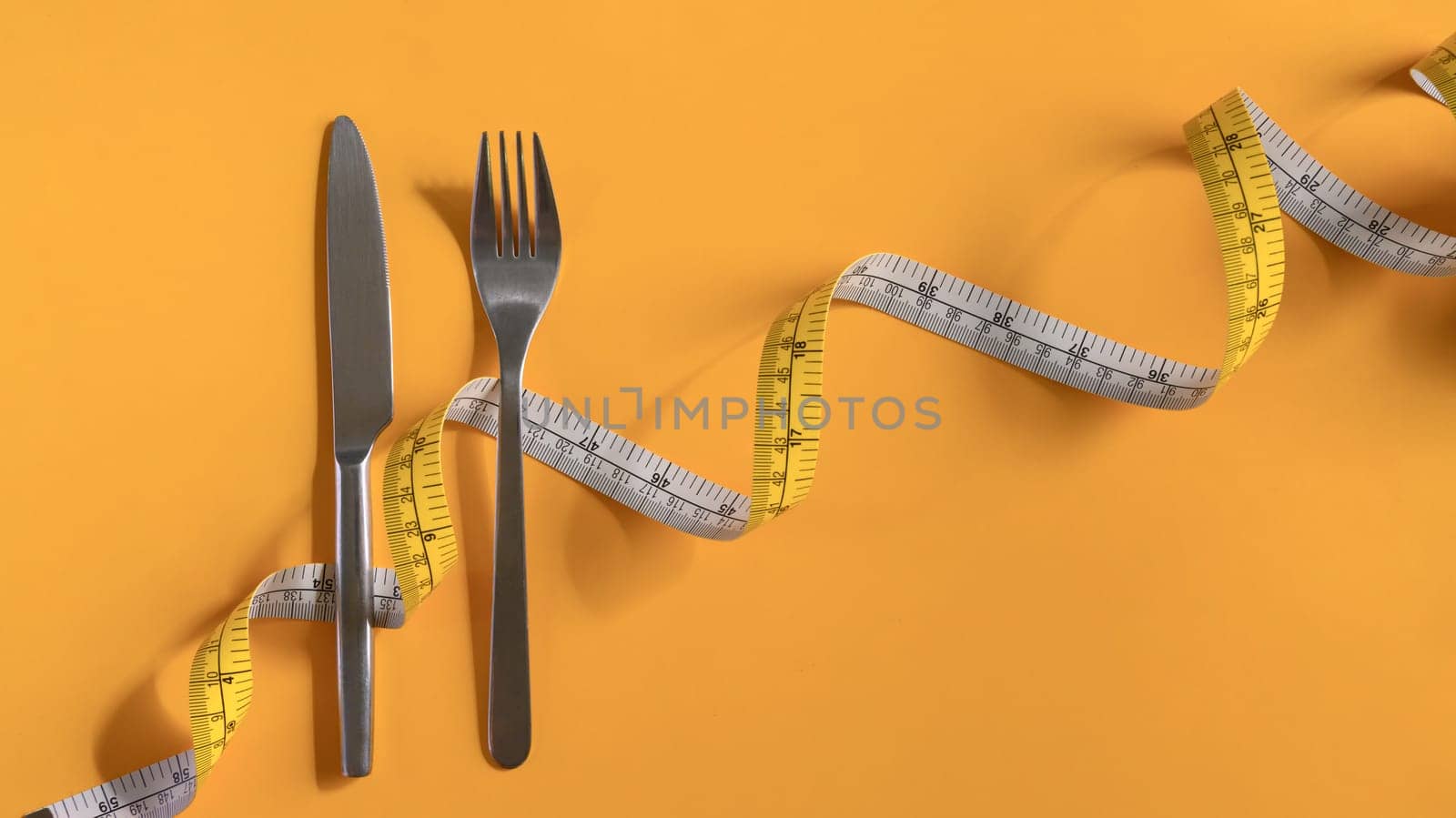 Fork and knife wrapped with measuring tape on yellow background. Weight loss and healthy concept.