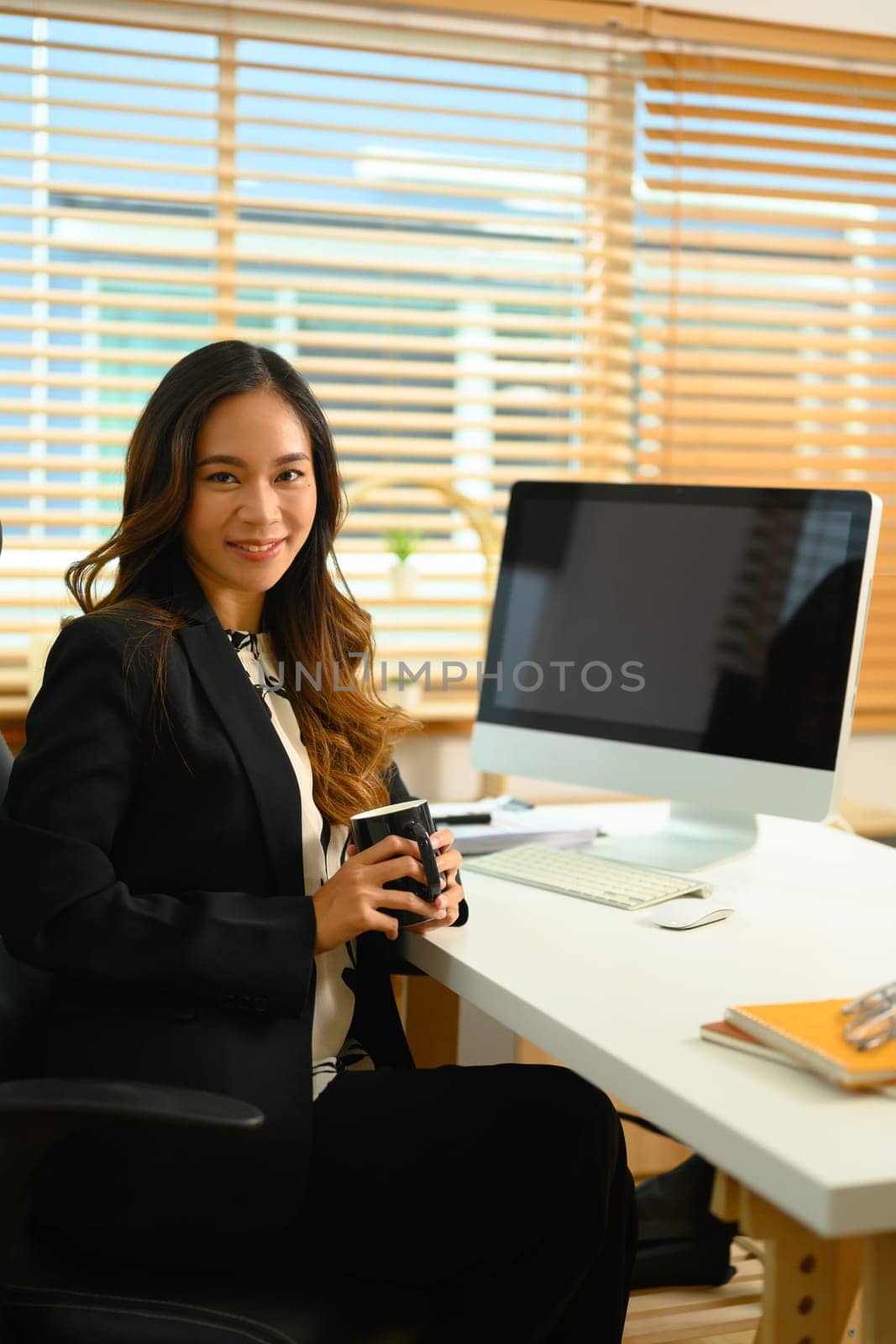 Portrait of successful businesswoman with cup of coffee sitting at desk and smiling at camera.