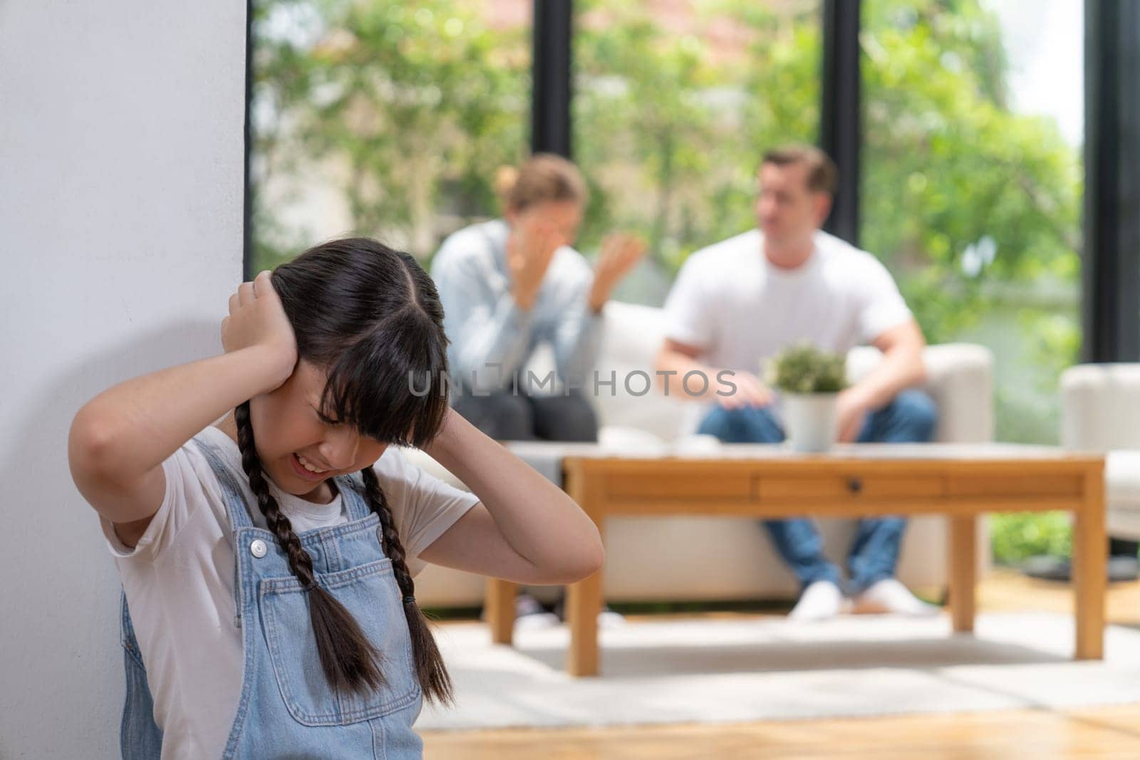 Stressed and unhappy girl huddle in corner, cover her ears with painful expression while her parent arguing in background. Domestic violence and traumatic childhood develop to depression. Synchronos