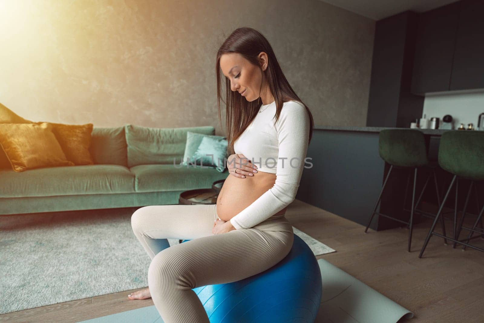 Calm pregnant woman sitting on a fitness ball and touching her belly. Happy mother doing supportive physical exercises for smooth pregnancy. by DariaKulkova