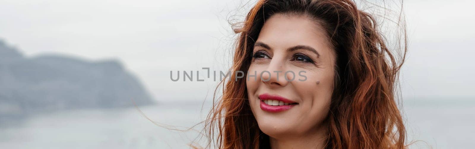 Portrait windswept hair happy woman against a backdrop of mountains and sea. Daylight illuminates the tranquil outdoor setting.