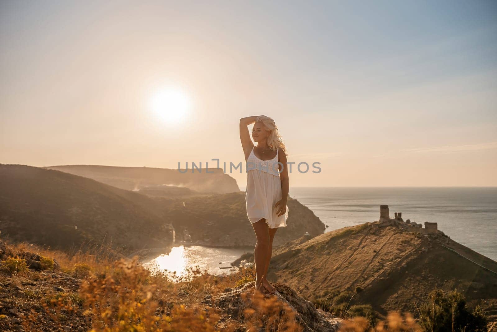 A blonde woman stands on a hill overlooking the ocean. She is wearing a white dress and she is enjoying the view