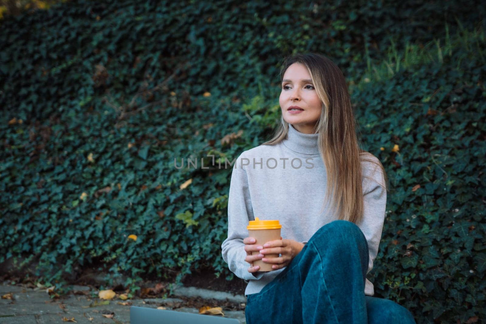 A woman sits on the ground with a cup in her hand. She is wearing a gray sweater and blue jeans. by Matiunina