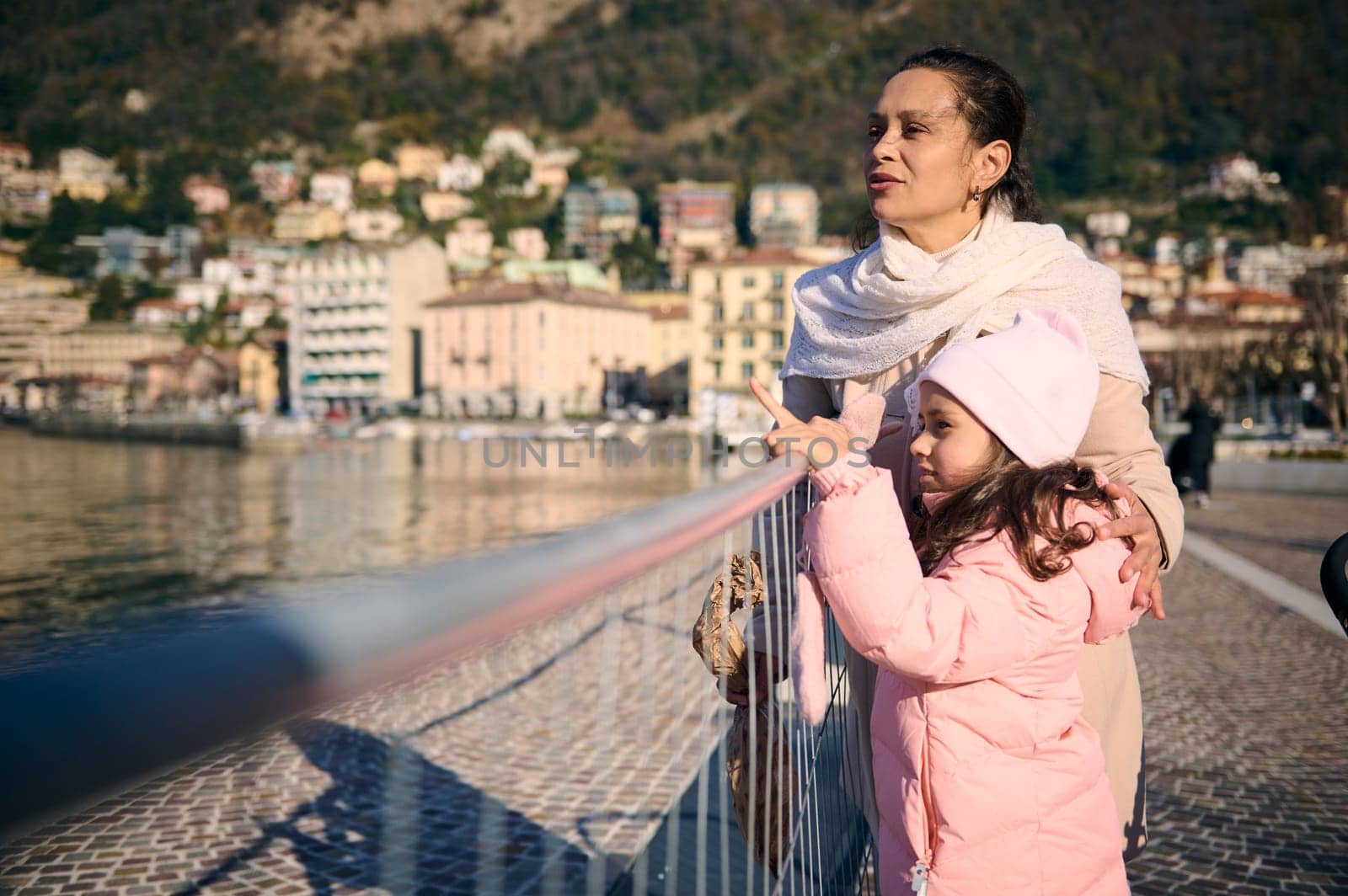 Young multi ethnic pretty woman and her cute daughter , a mother and little child girl in warm winter clothes, standing together on the promenade, admiring the beauty of the lake of Como in Italy