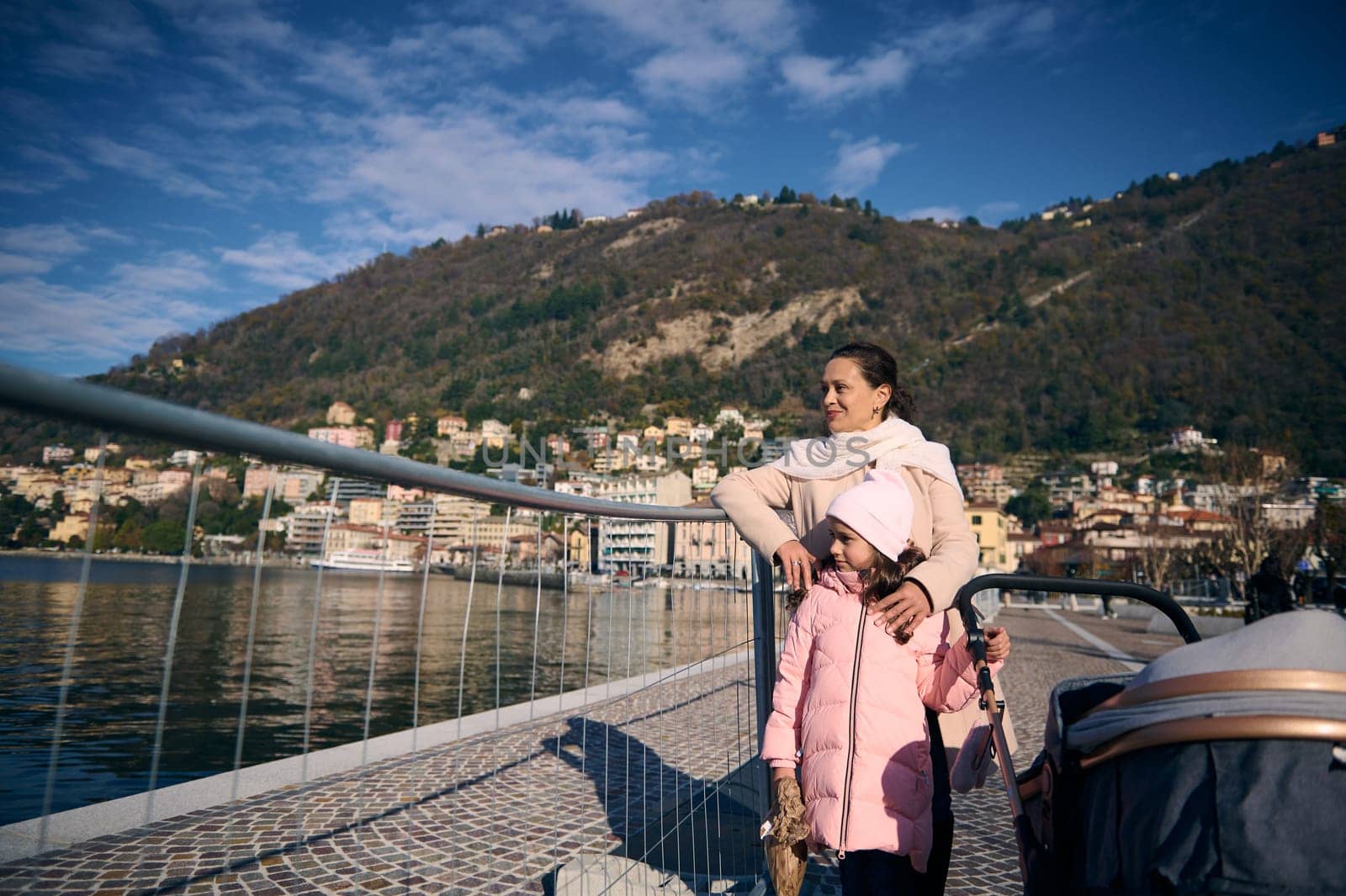 Beautiful smiling woman and her little kid, cute daughter walking on the promenade near the lake of Como. Stylish baby pram in the foreground. Family. Mother and children enjoying family walk outdoors