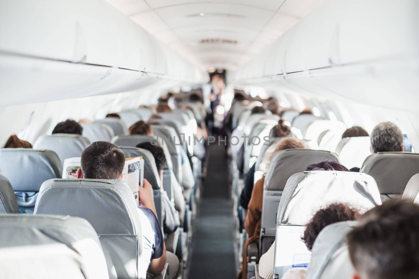 Interior of airplane with passengers on seats and stewardess in uniform walking the aisle, serving people. Commercial economy flight service concept