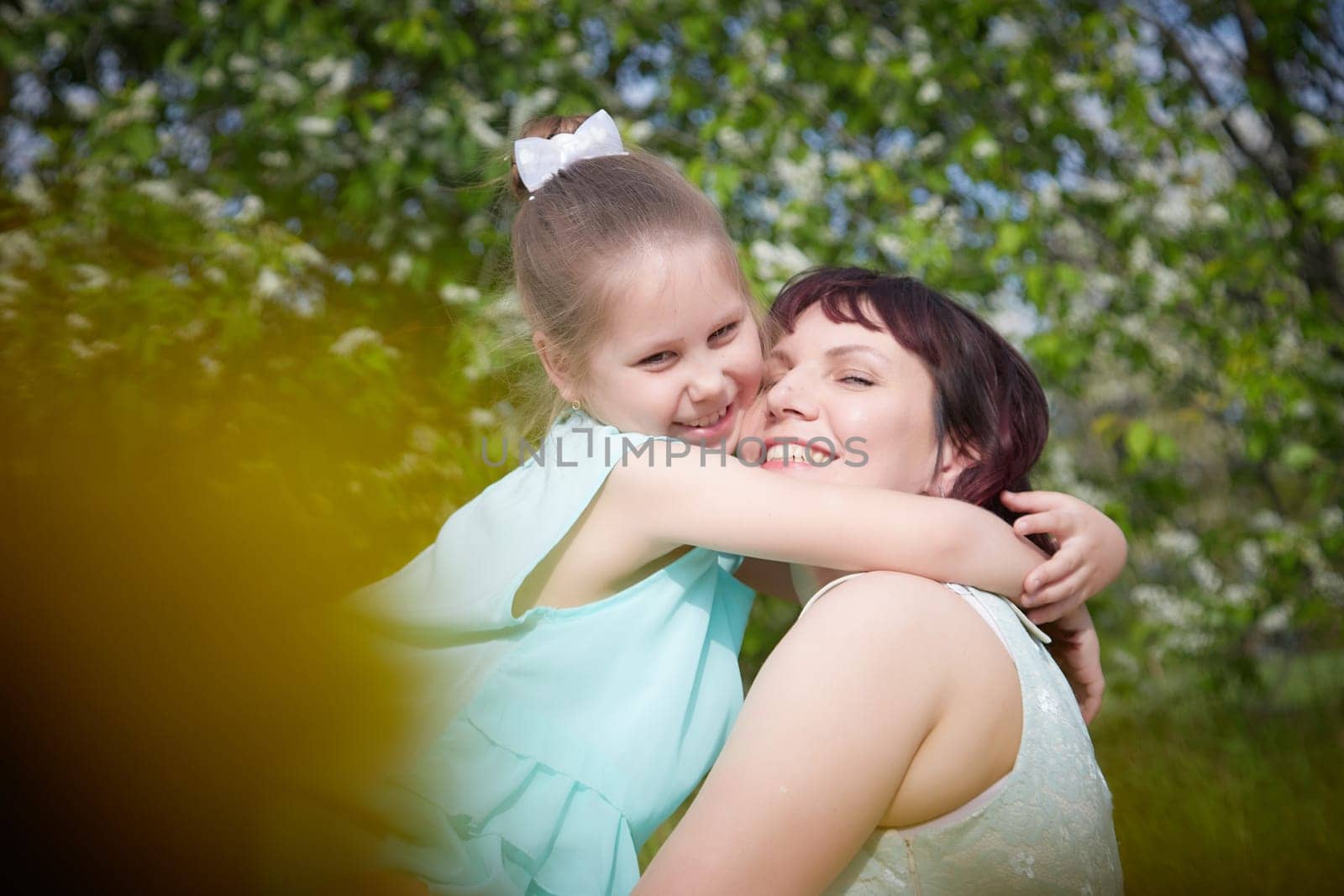 Happy mother and daughter enjoying rest, playing and fun on nature on a green lawn with dandelions and blooming apple tree on background. Woman and girl resting outdoors in summer and spring day