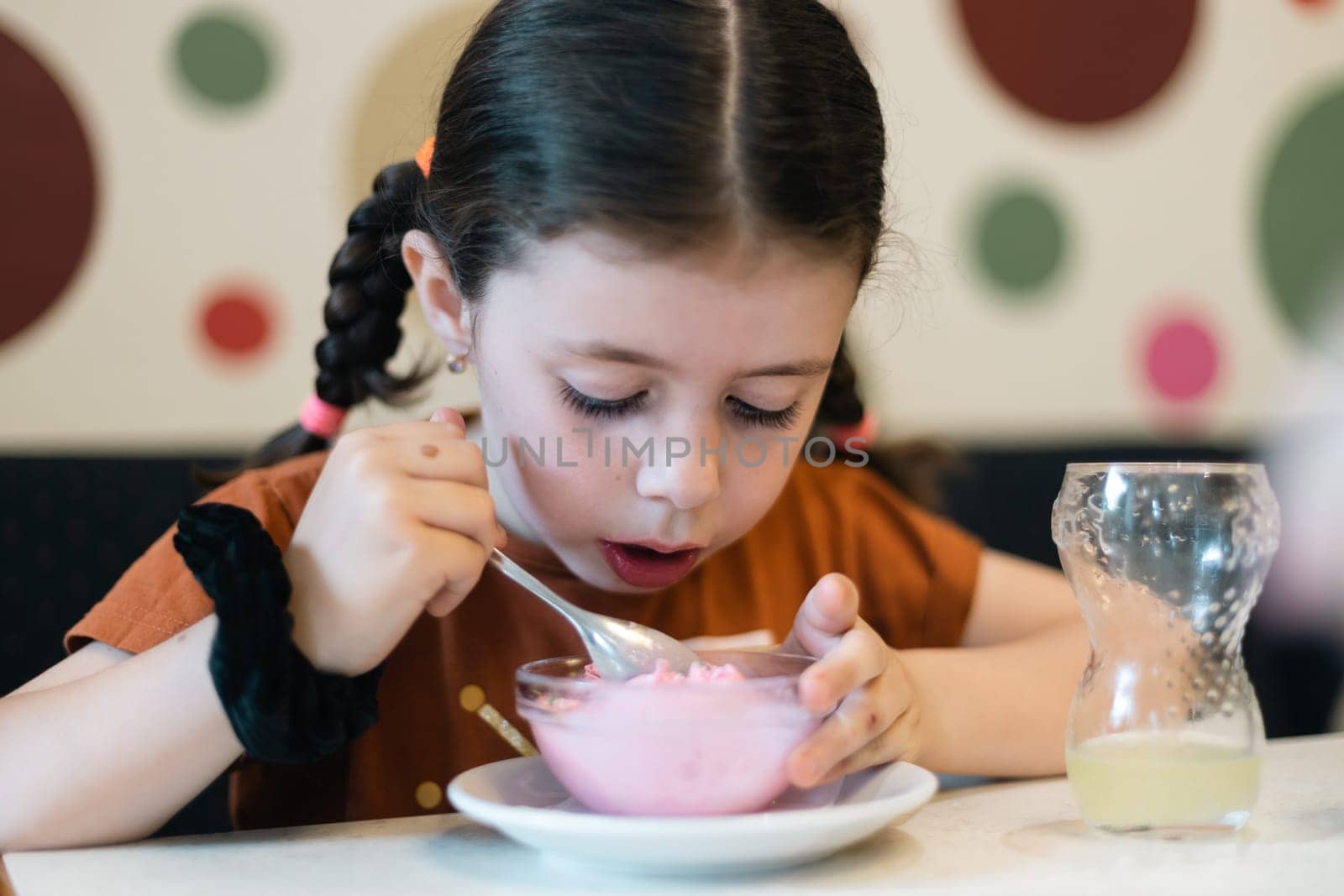 Portrait of one beautiful caucasian brunette girl looking with surprise and eating ice cream while sitting on a sofa in a cafe against the background of an abstract wall, close-up side view.