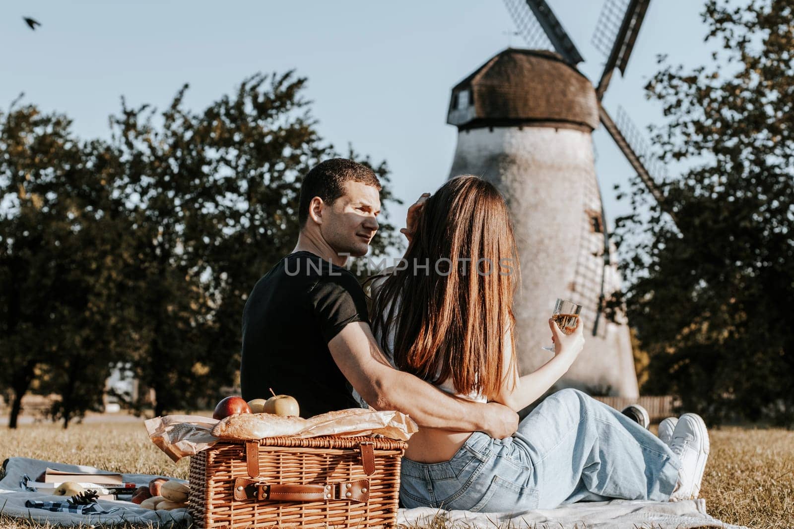 One beautiful caucasian young couple sit from the back in an embrace tenderly looking at each other on a bedspread with a wicker basket and fruits in the park against the backdrop of an old mill, side view close-up.