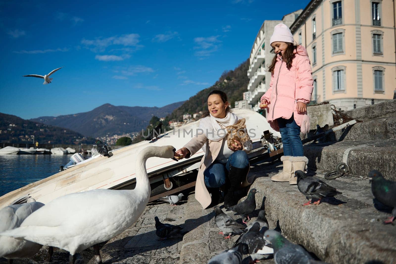People, nature and animals. Happy mother and daughter feeding swans and ducks at the lake of Como in Italy, on a sunny cold winter day. Beautiful mountains and clean clear blue sly on the background.