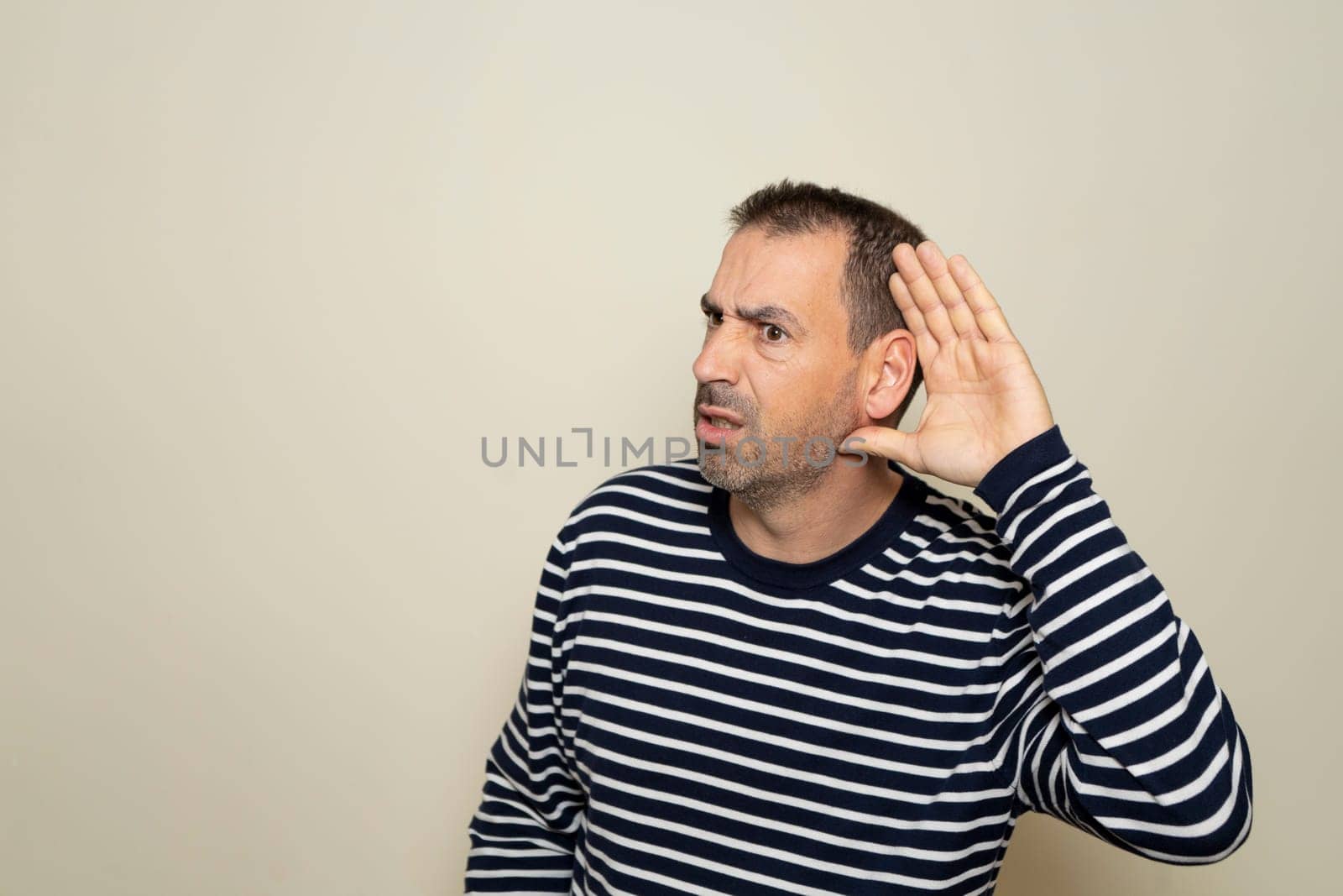 Hispanic man with beard in his 40s wearing a striped sweater listening to rumors keeping hand near ear isolated over beige color background. by Barriolo82