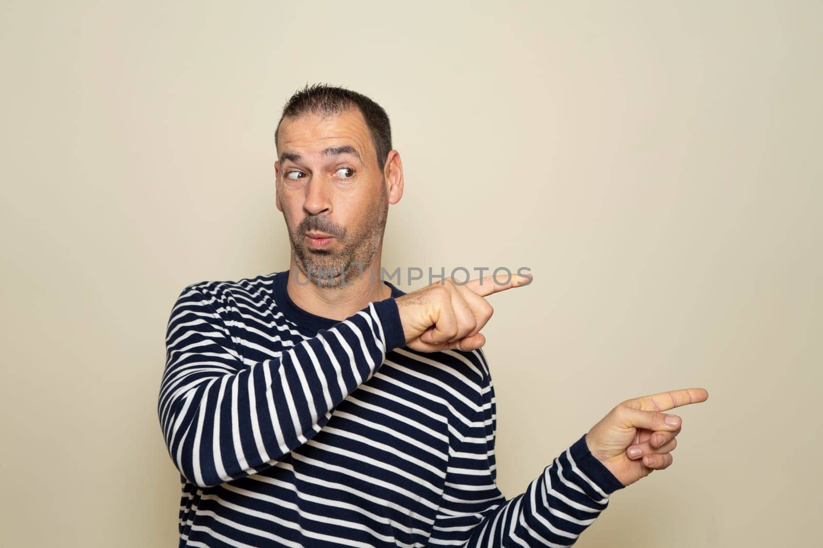 Bearded Hispanic man in his 40s wearing a striped sweater pointing to the side with his index fingers with an expression of surprise. Isolated on beige background. by Barriolo82