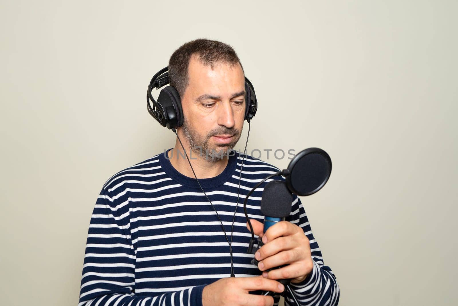 Bearded Hispanic man in his 40s wearing a striped sweater looking confused while inspecting a microphone and headphones, isolated on beige background. by Barriolo82