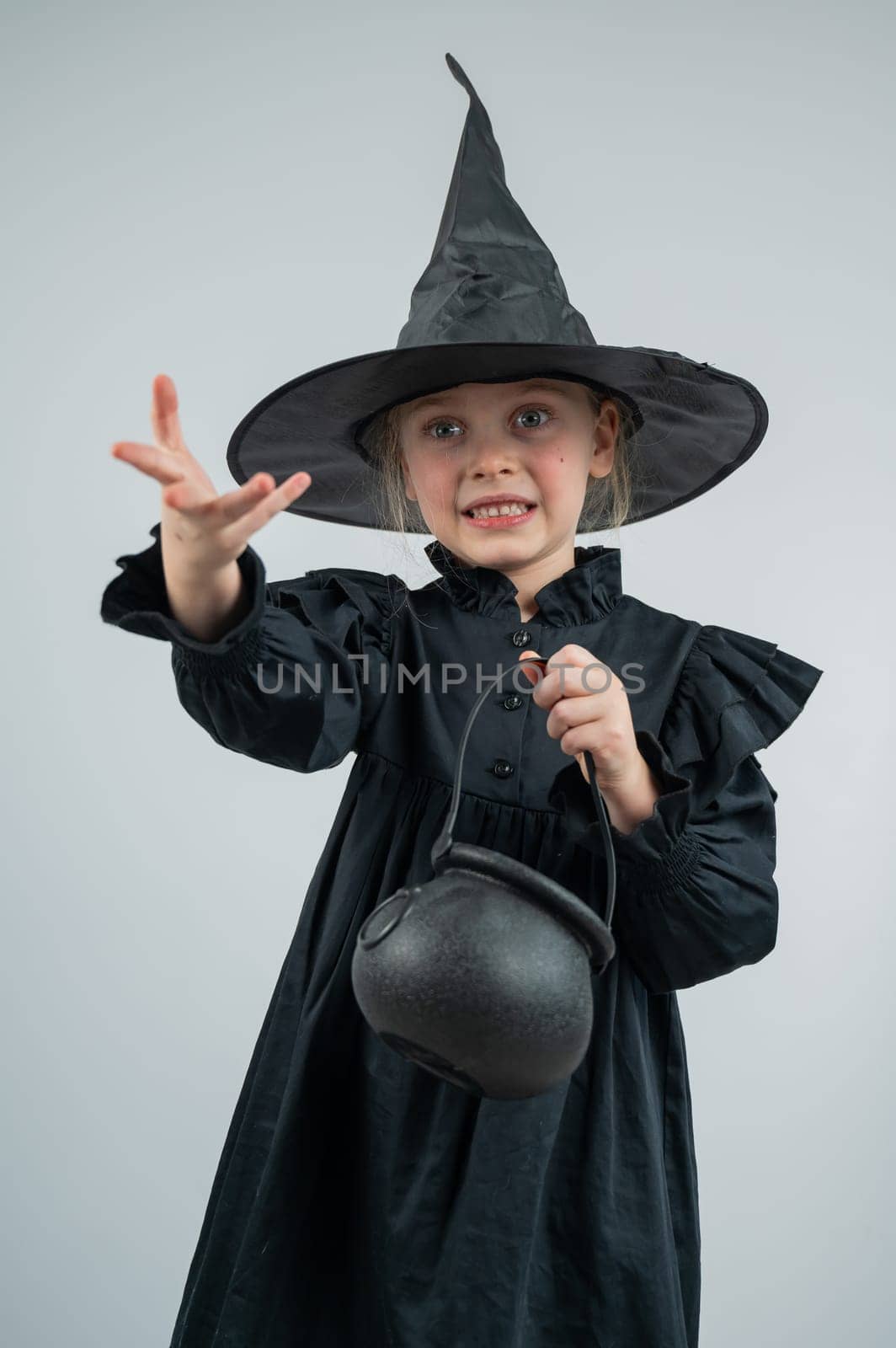 Portrait of a little Caucasian girl in a witch costume holding a cauldron on a white background. Vertical photo. by mrwed54