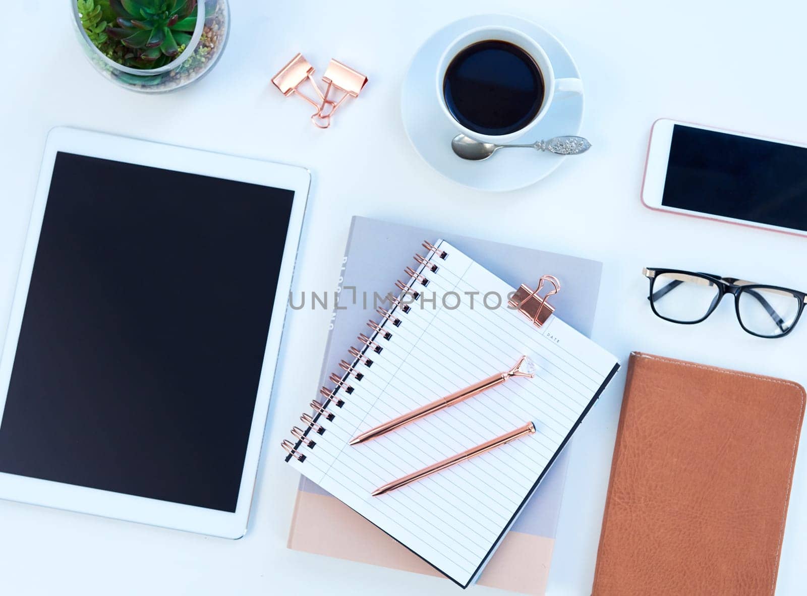 Tablet, notebook and coffee on desk in studio for working, writing notes and planning for career. Creative workplace, business and diary, digital tech and stationery on blue background for job.