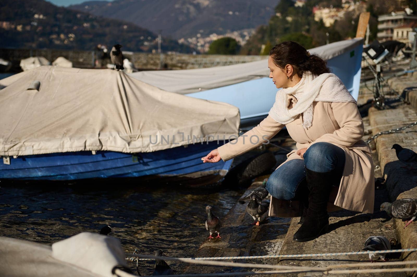 Young beautiful woman crouching by ducks and swans on the lake of Como, feeding birds with bread, relaxing on the lakefront on a sunny and cold winter day. The province of Milan, Como, Lombardy, Italy by artgf