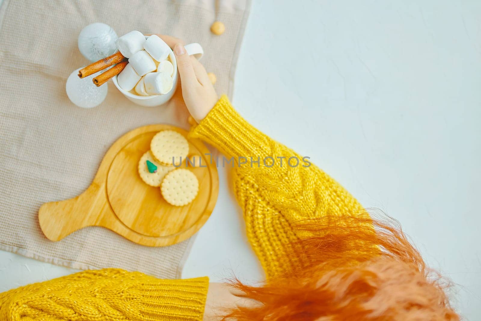 Woman in a yellow sweater drinks hot cocoa with marshmallow and cinnamon rolls in a white mug and cookies and a figure of a Christmas tree