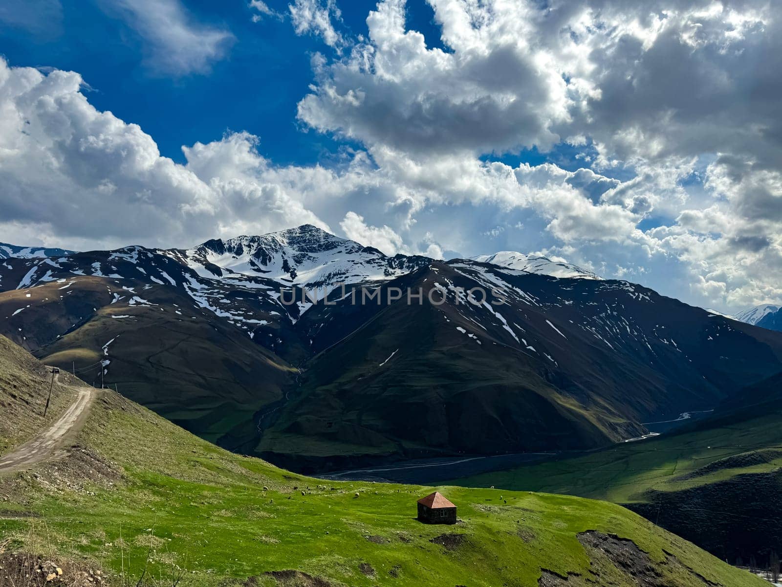 Solitary house in verdant valley with snow capped mountains under dynamic sky with billowing clouds, depicting isolation and beauty of nature. High quality photo