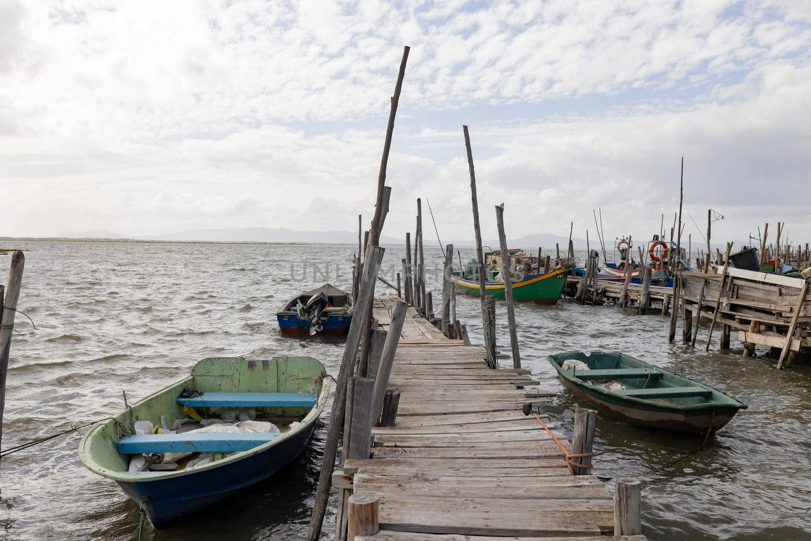 A pier with several boats docked at it. The boats are of various sizes and colors, including a green and blue boat. The pier is located near the water, and the boats are tied to the dock