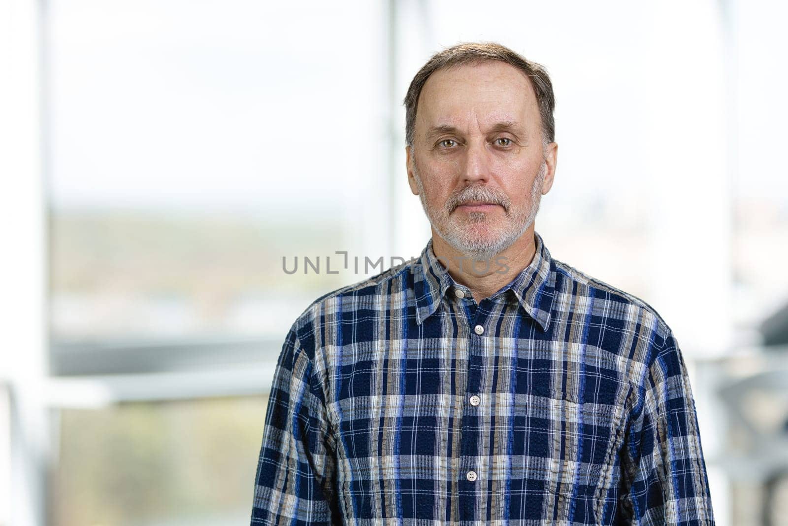 Portrait of a handsome mature man in office. Blue casual checkered shirt. Blurred window background.