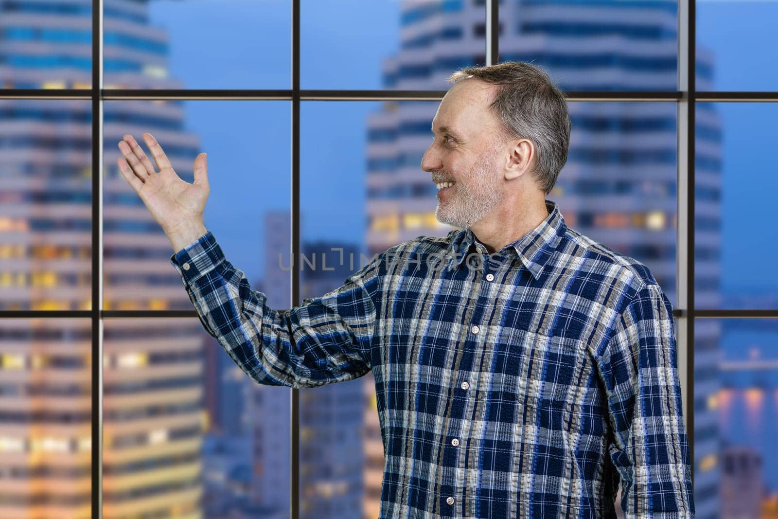 Handsome cheerful mature senior man presenting copy space with his hand. Checkered windows with night cityscape background.