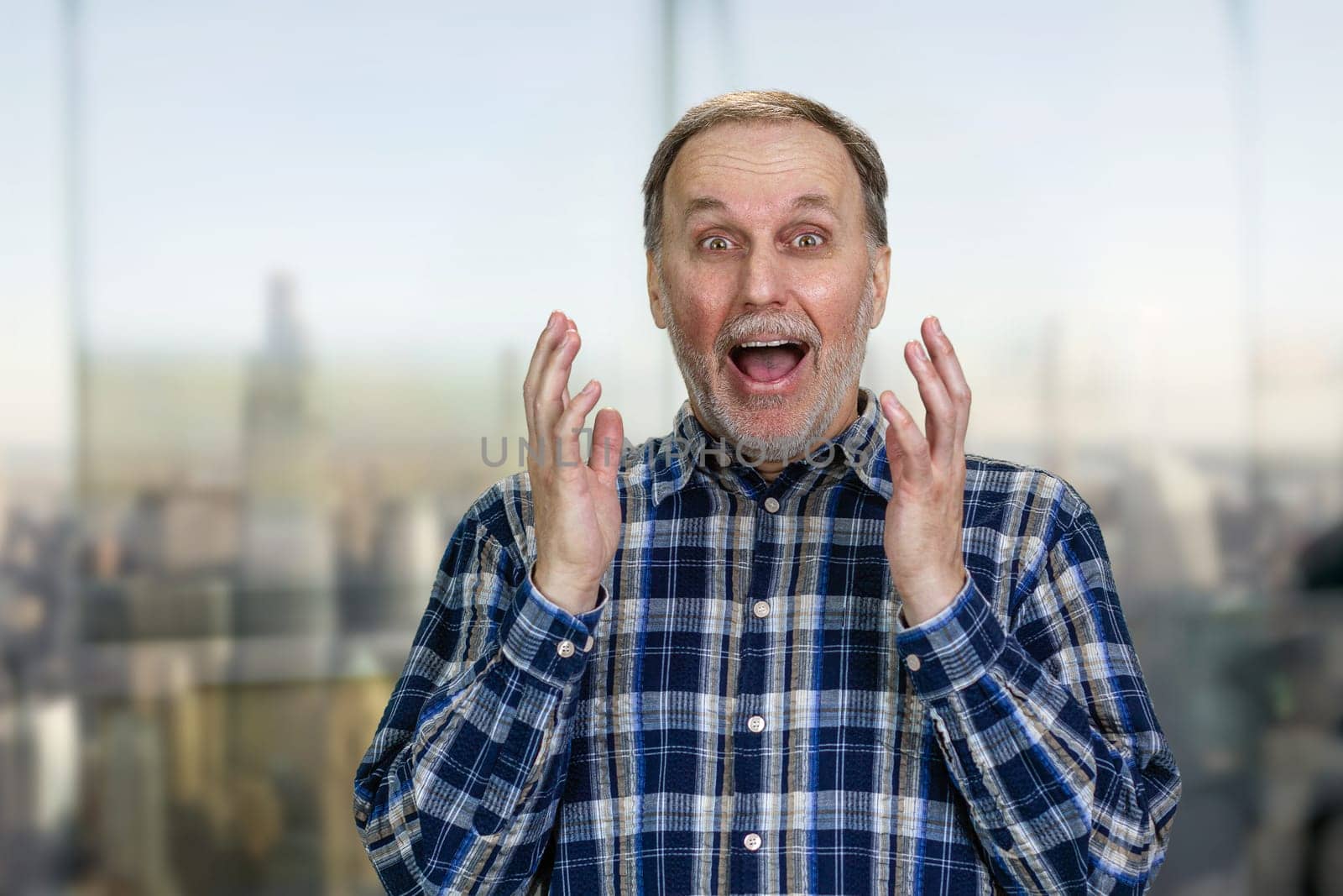 Handsome excited senior manture man standing indoors. Surprised man with mouth open. Blurred cityscape background.