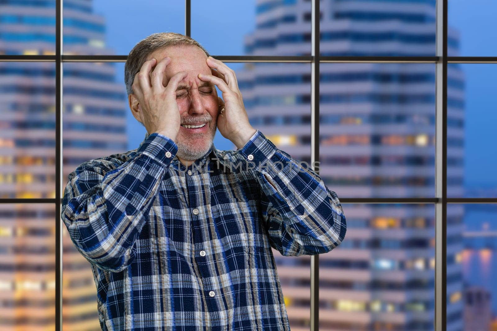 Handsome mature man holds his head with both hands. Checkered windows background with night city view.