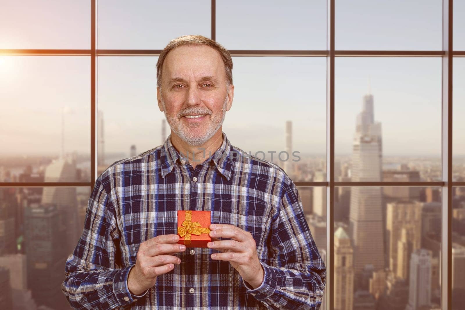 Happy smiling handsome senior man is holding a red gift box in both hands. Evening cityscape background.