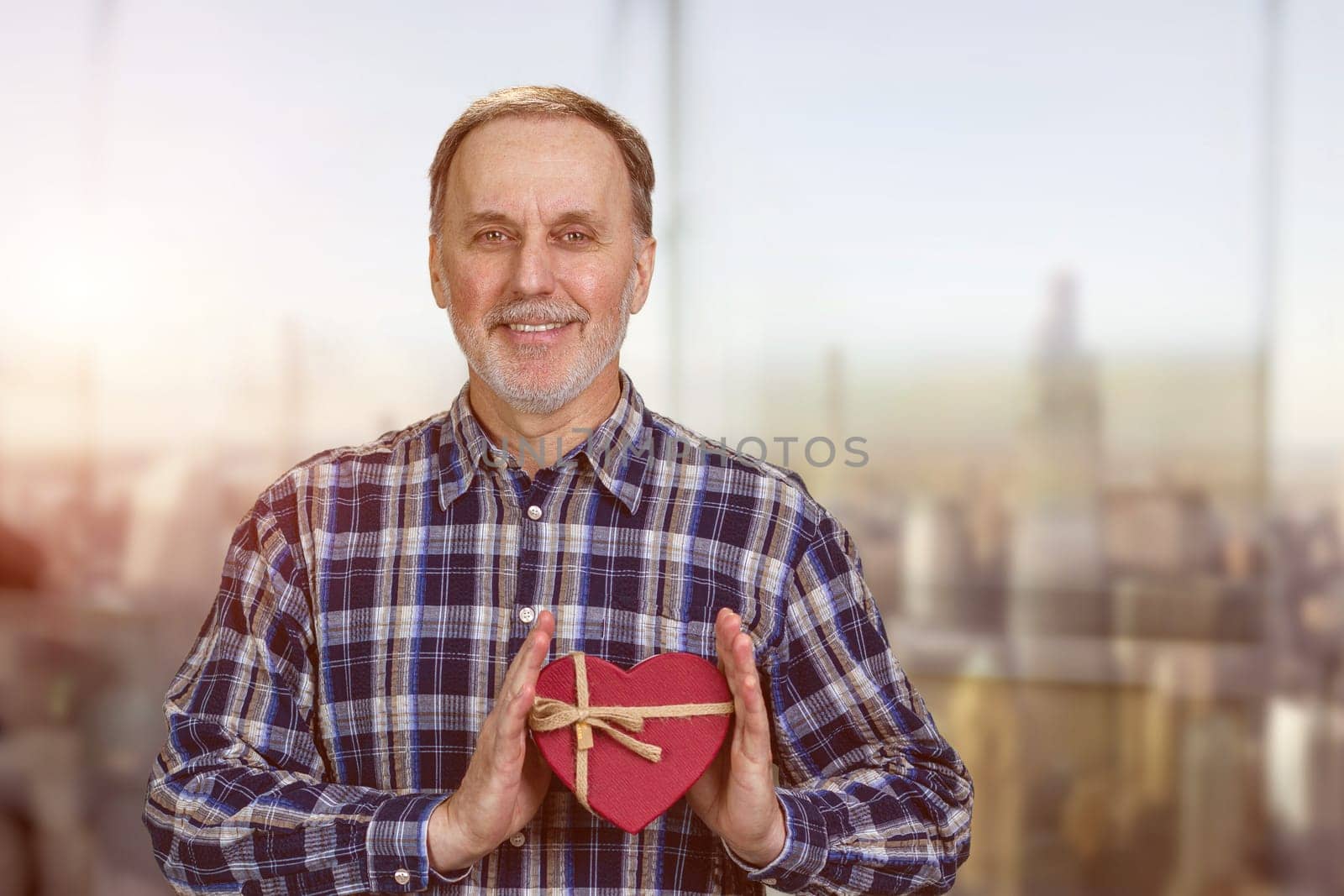 Happy smiling handsome senior man is holding a heart shape gift box. Blurred cityscape background.