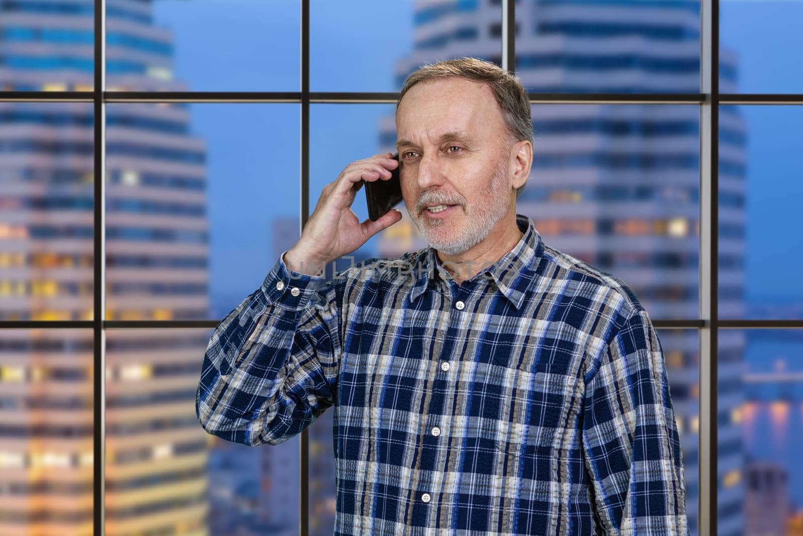 Handsome mature businessman is talking on the mobile phone standing in office. Checkered window background with view on the night city.