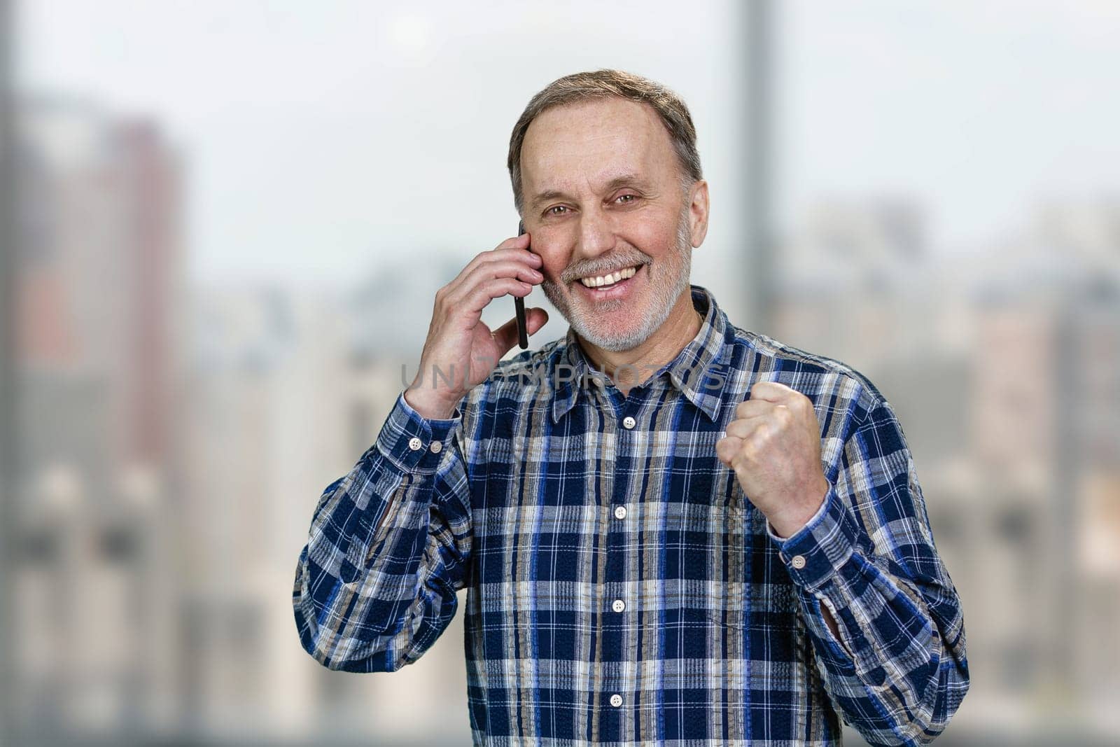 Happy cheerful senior man is talking on phone celebrating success. Blurred urban scape background.