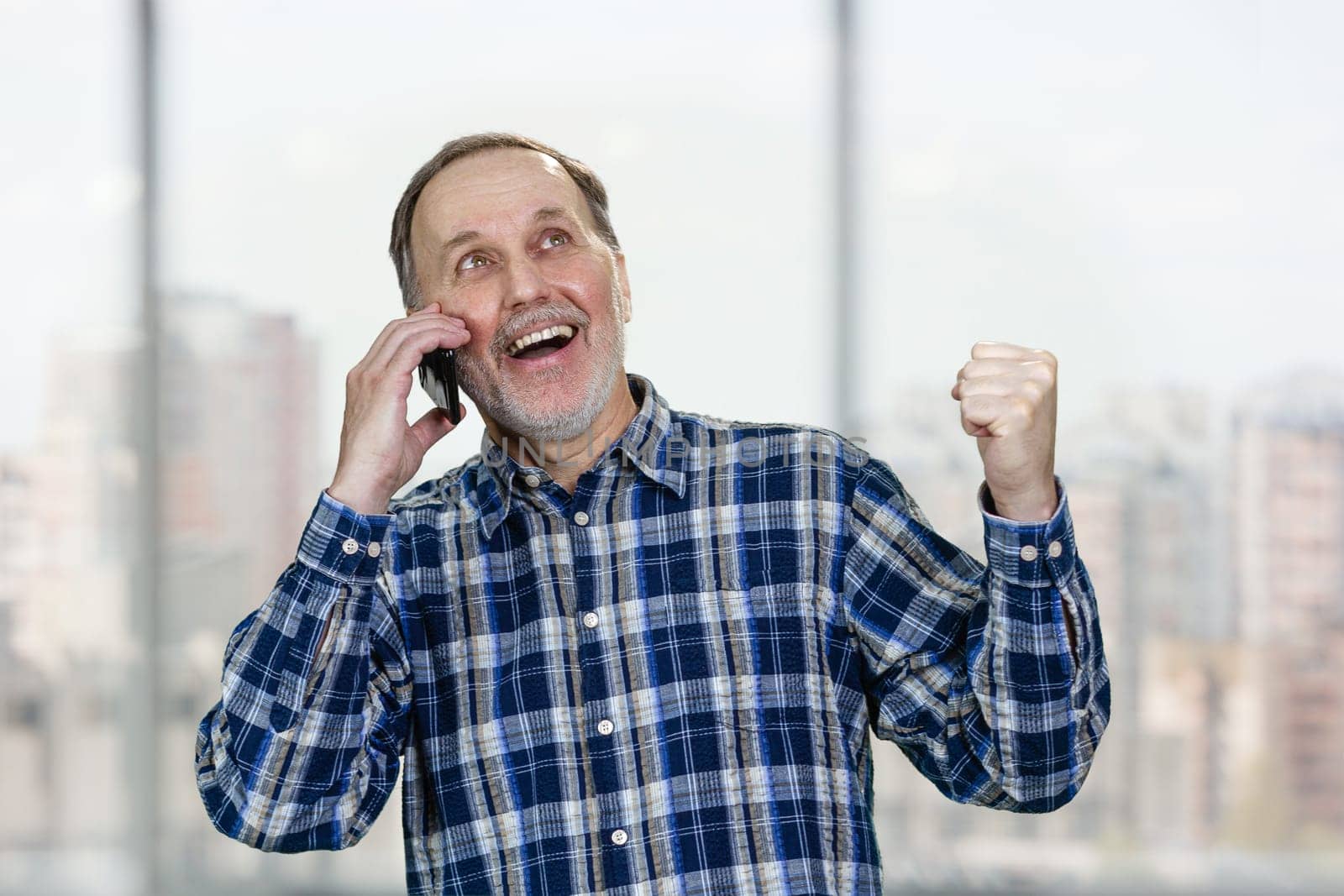 Happy excited senior man is talking on phone celebrating success. Blurred urban scape background.