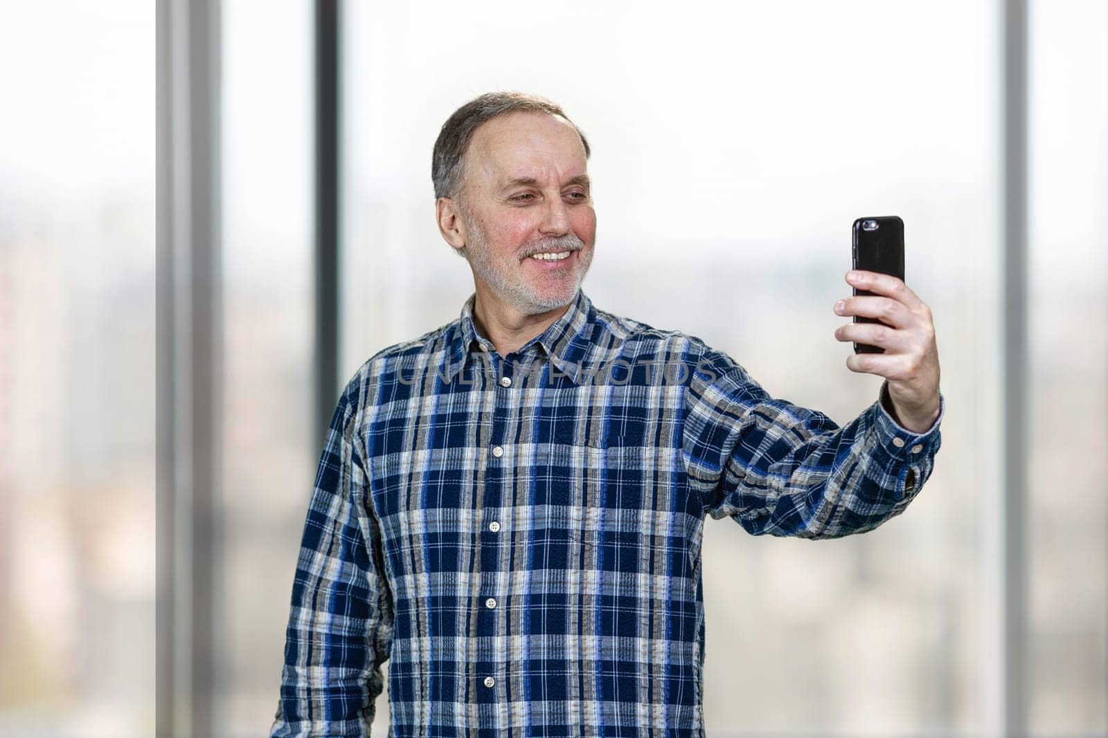 Portrait of smiling mature man taking a selfie on his smartphone camera. Standing indoors on windows backgorund.