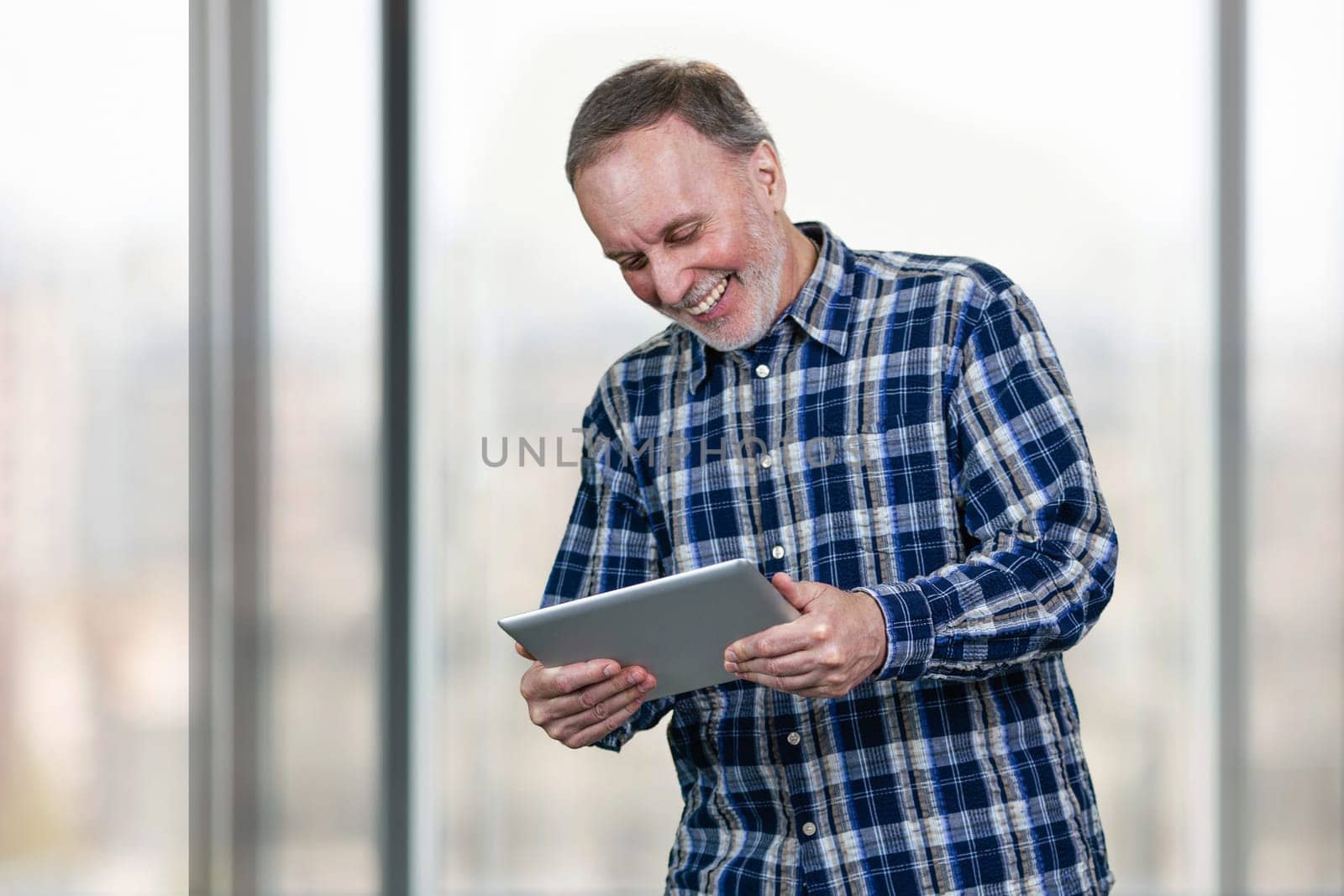 Smiling mature man holding tablet pc in both hands. Huge office windows background.