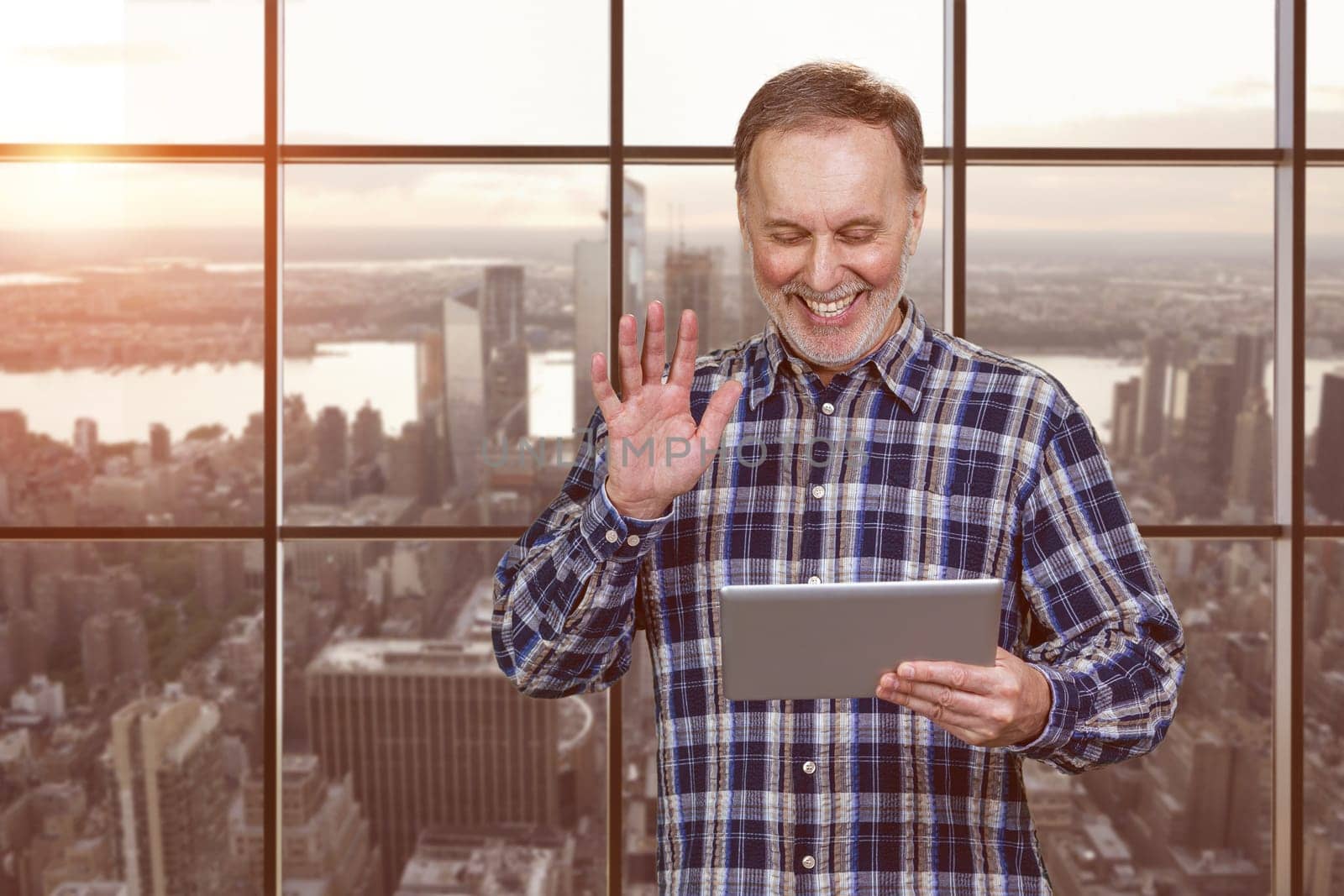 Portrait of happy mature man waving his hand to someone online. Checkered windows background with cityscape view.