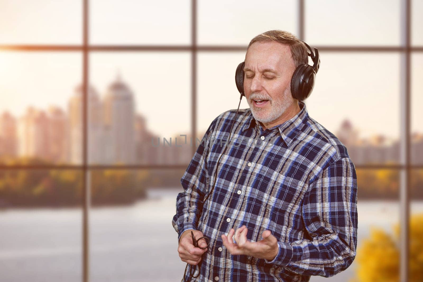Portrait of mature man listening to music in headphones. Checkered windows background with city river view.