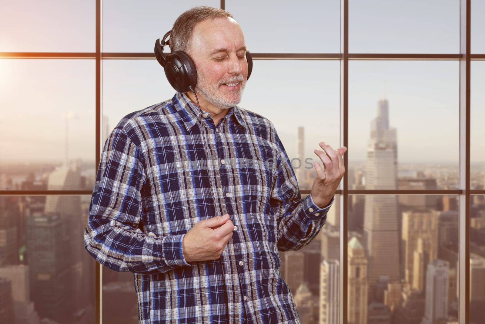 Mature man listening to music in headphones and enjoying. Checkered window background with cityscape view.