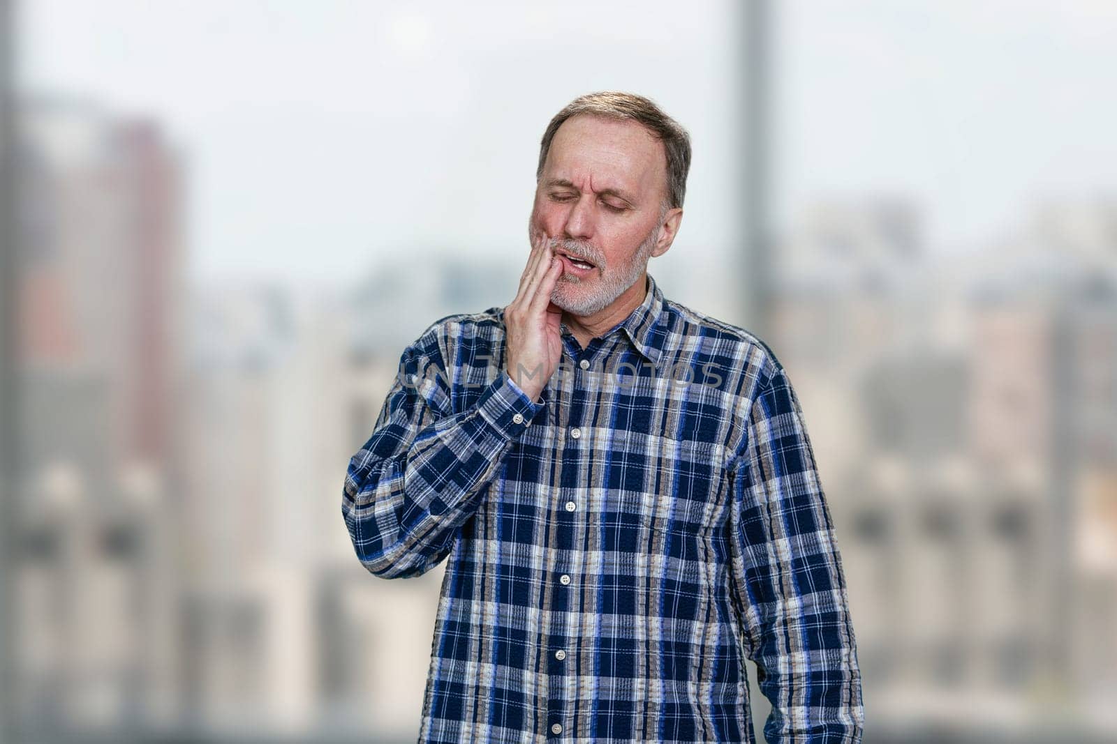 Mature office worker in checkered shirt having toothache. Blurred windows background.