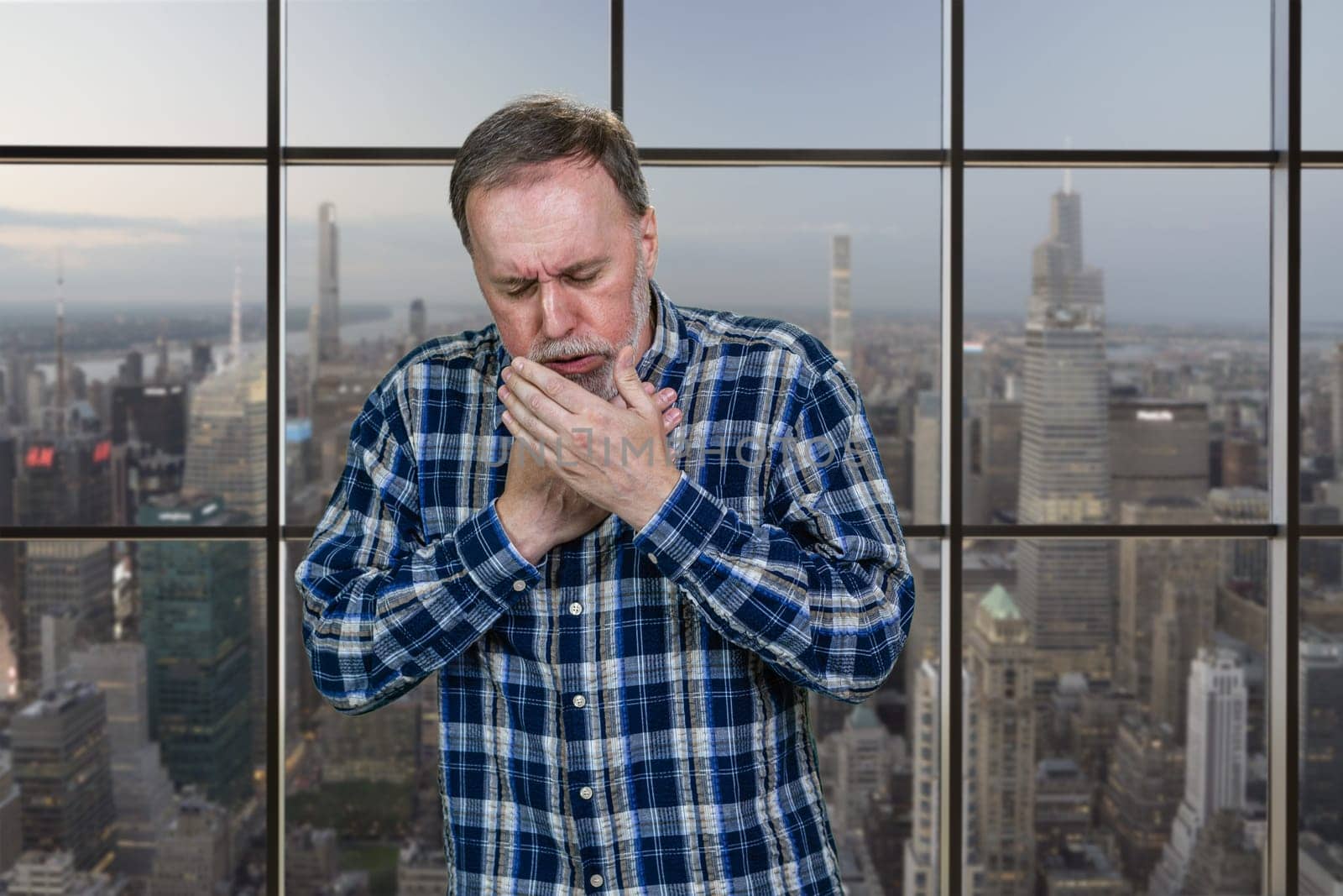 Sad mature caucasian man is coughing standing indoors. Windows background with cityscape view.