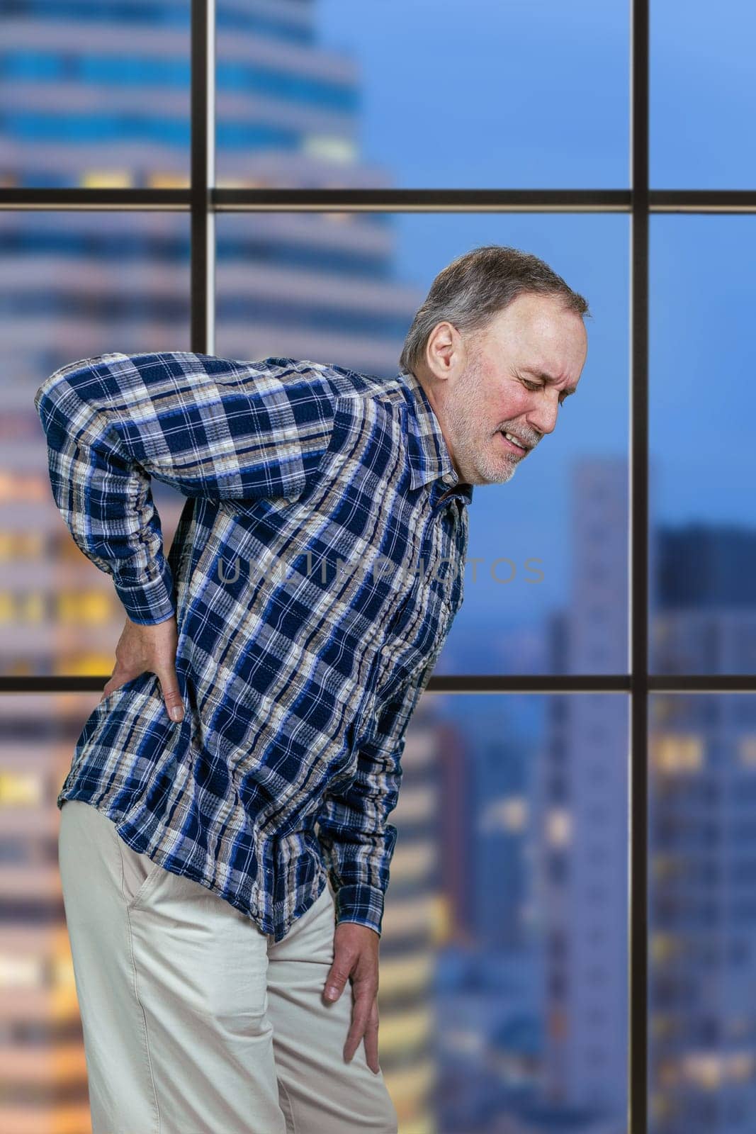 Aged mature man is having a back pain touching his back. Vertical shot of an old casual man suffering from backache.