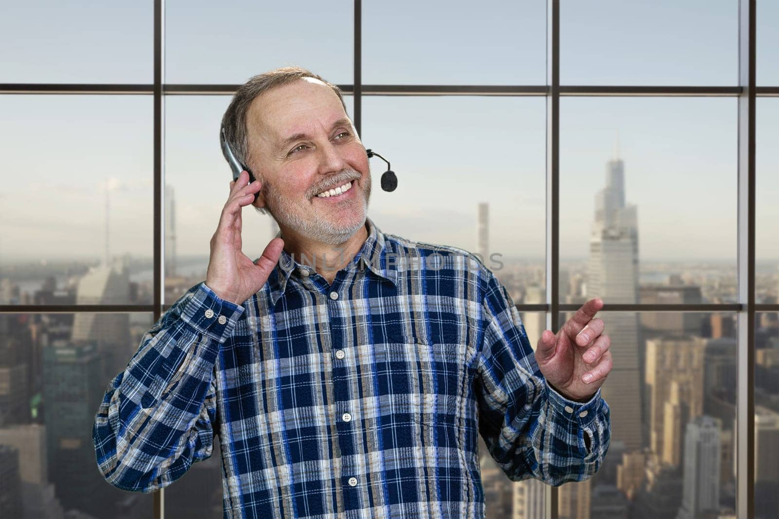 Portrait of smiling mature male call center customer support worker. Checkered window backgroud with cityscape view.