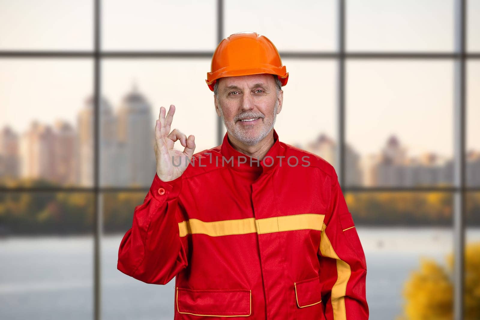 Happy smiling construction worker in red uniform shows okay gesture sign. Checkered window background.