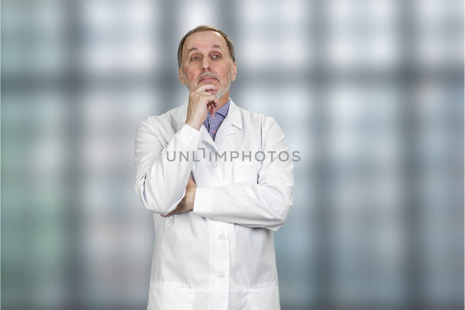Thoughtful pensive senior male doctor looking away. Blurred checkered window background.