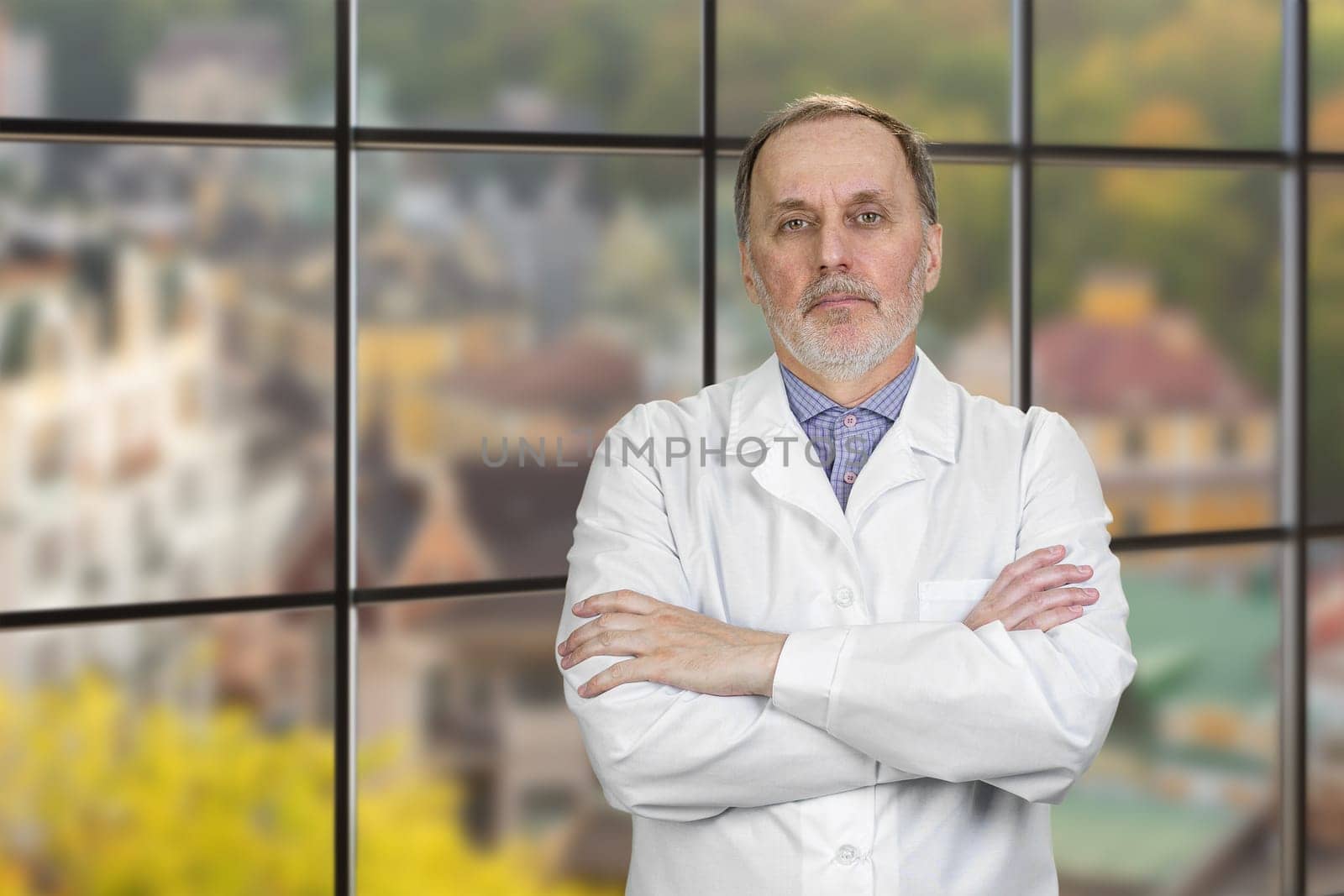 Portrait Of Happy Mature Male Doctor With Folded Arms. Checkered windows backgorund with cityscape view.