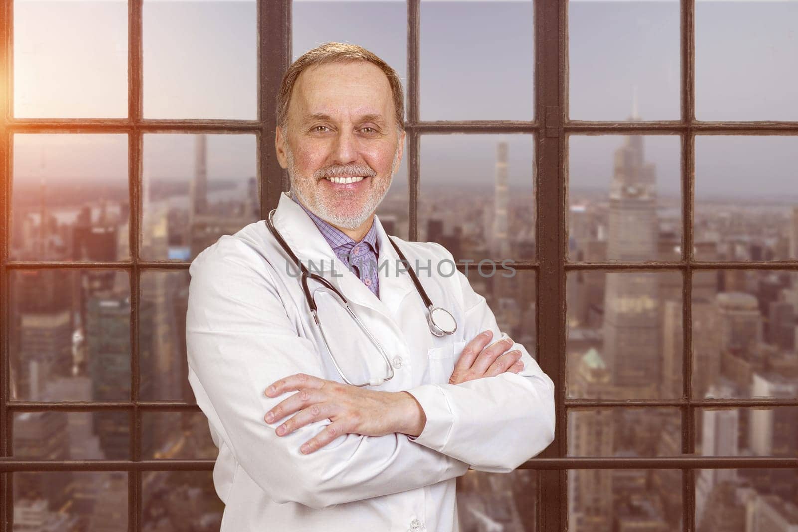 Portrait of happy senior doctor with folded arms. Evening cityscape view.