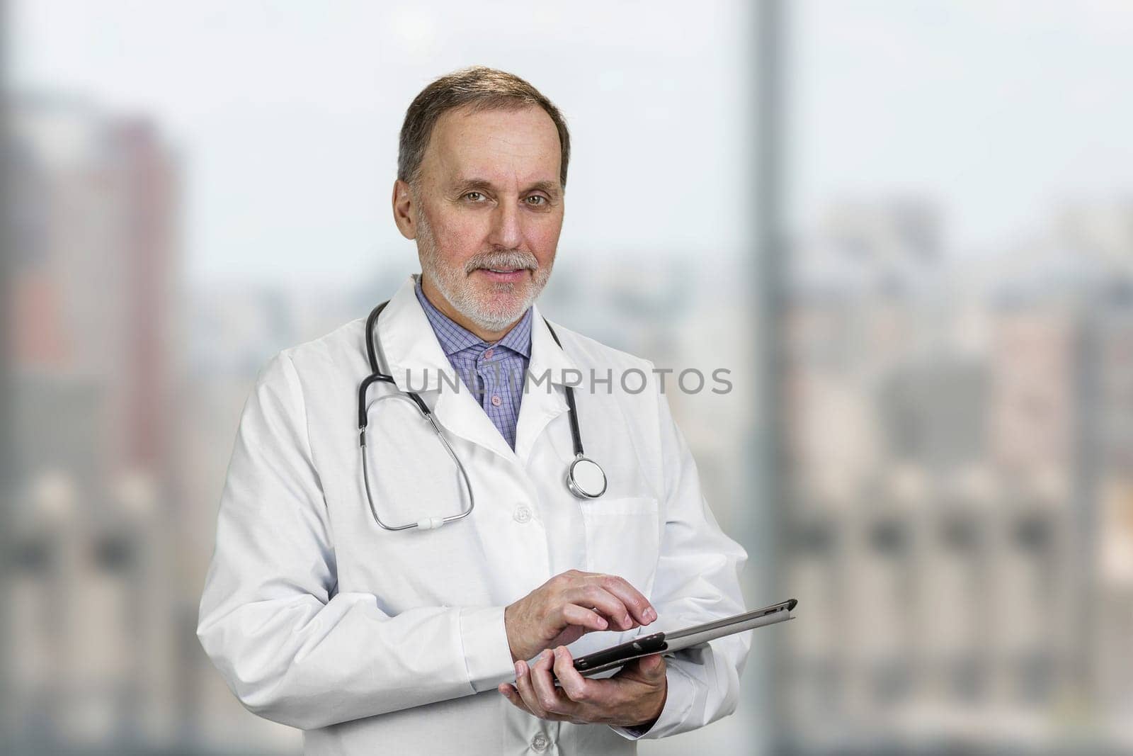 Portrait of mature senior male doctor holding digital tablet standing indoors. by super_picture