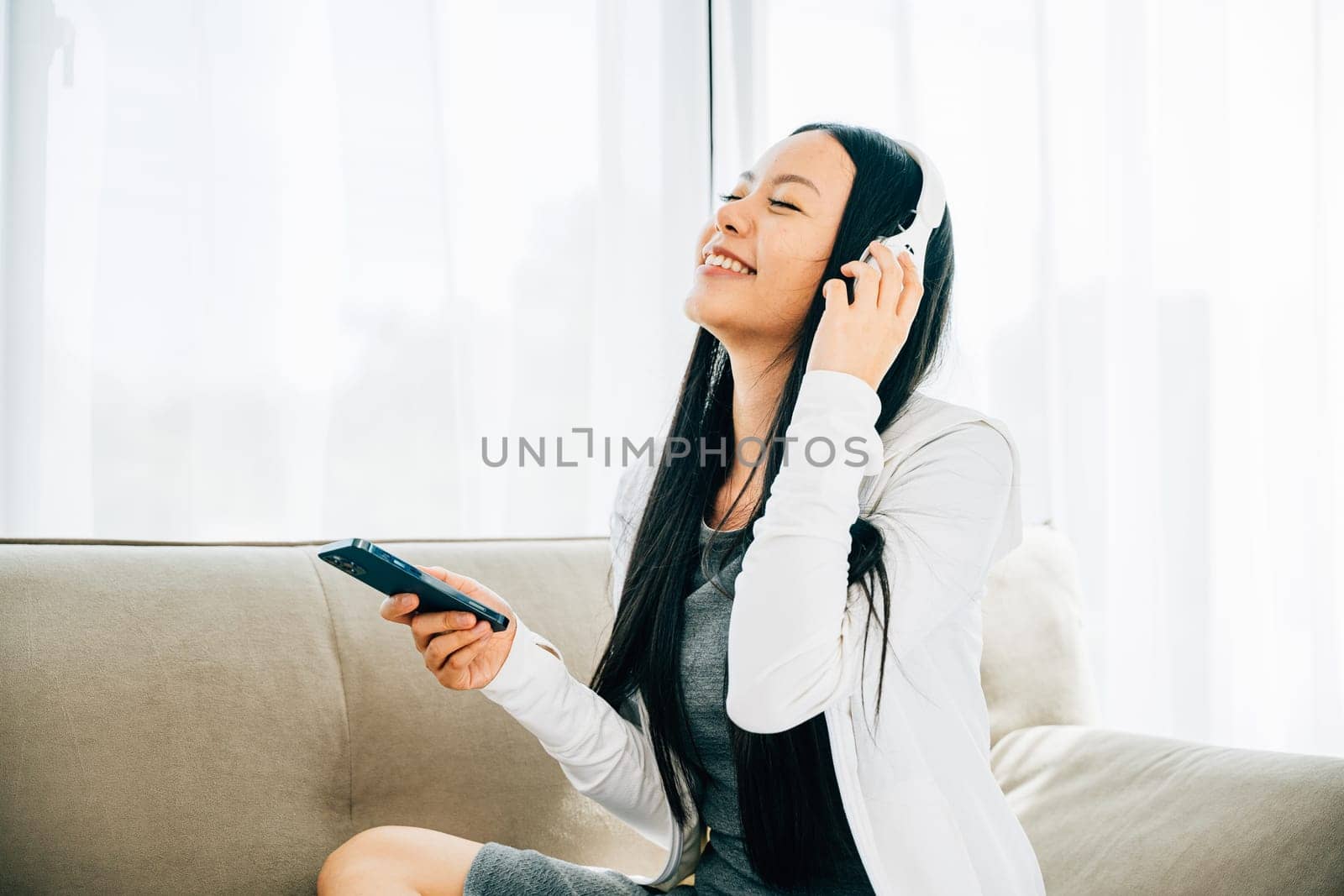 Woman with smartphone and headphones relaxes on sofa listens to music by Sorapop